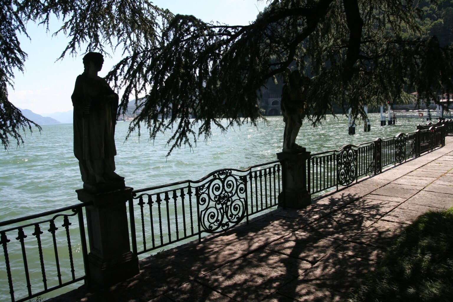 Scenic view of a lakeside promenade featuring a decorative iron fence, a statue, and trees casting shadows over the path, with calm waters in the background. | Only on SopranoVillas