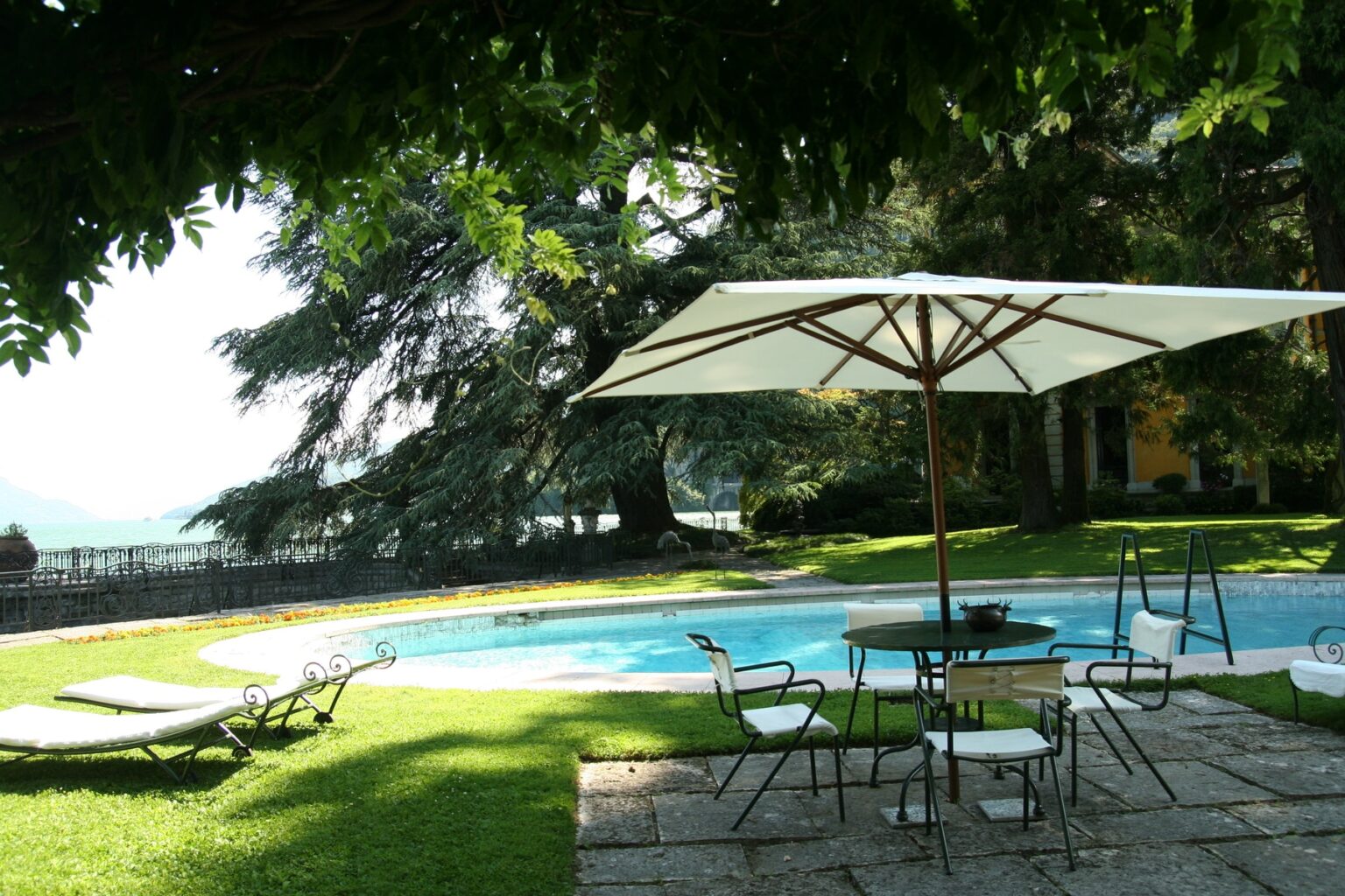 Scenic outdoor pool area featuring lounge chairs, a shaded table with chairs, and lush greenery, ideal for relaxation and leisure. | Only on SopranoVillas