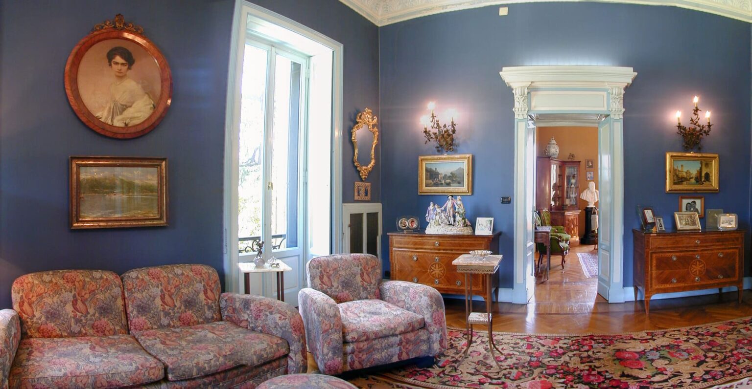 Cozy living room featuring floral-patterned sofas, blue walls adorned with vintage portraits and paintings, and elegant wooden furniture. The room includes a decorative chandelier and opens to a bright doorway leading to another area of the house. | Only on SopranoVillas