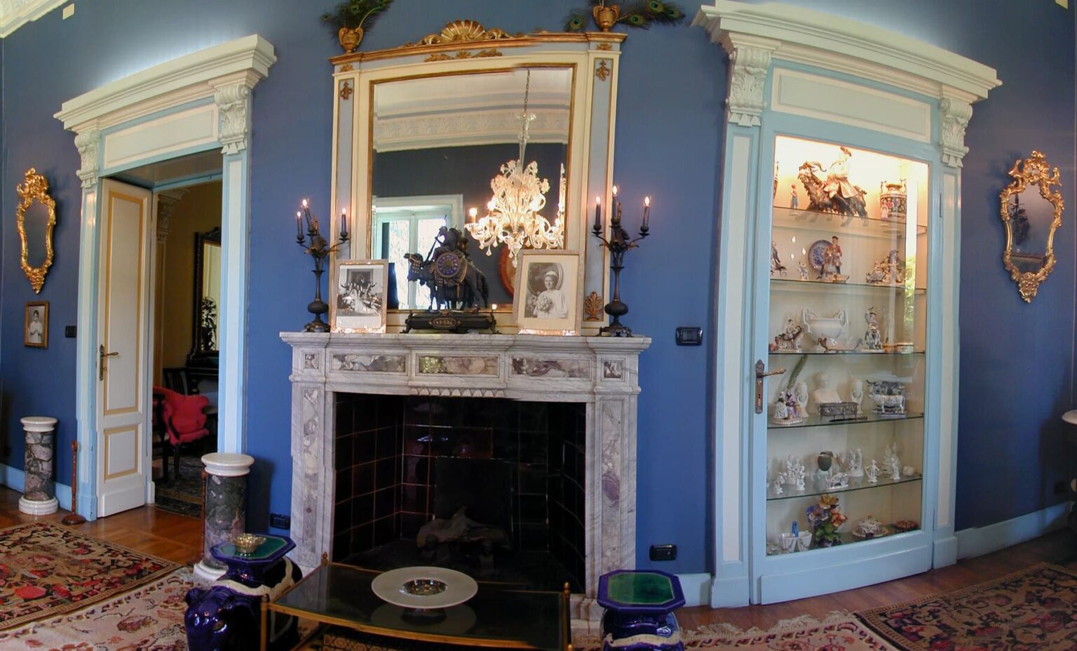 Elegant living room featuring a marble fireplace, ornate mirror, and decorative shelves filled with porcelain collectibles. The walls are painted in a rich blue, complemented by antique furniture and a chandelier, creating a sophisticated atmosphere. | Only on SopranoVillas