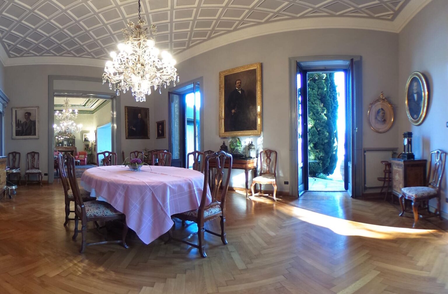 Elegant dining room featuring a round table set with a pink tablecloth, surrounded by wooden chairs. The room is illuminated by a crystal chandelier and showcases classic artwork on the walls. Large doors open to a garden, enhancing the spacious and inviting atmosphere. | Only on SopranoVillas