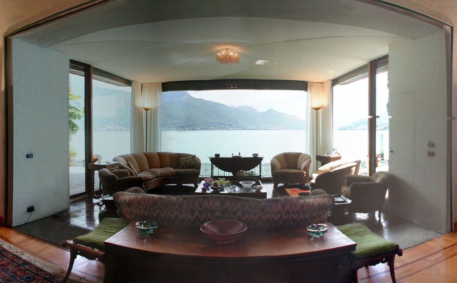 Cozy living room with large windows showcasing a scenic lake view, featuring elegant furniture, a wooden table with decorative items, and soft lighting. | Only on SopranoVillas