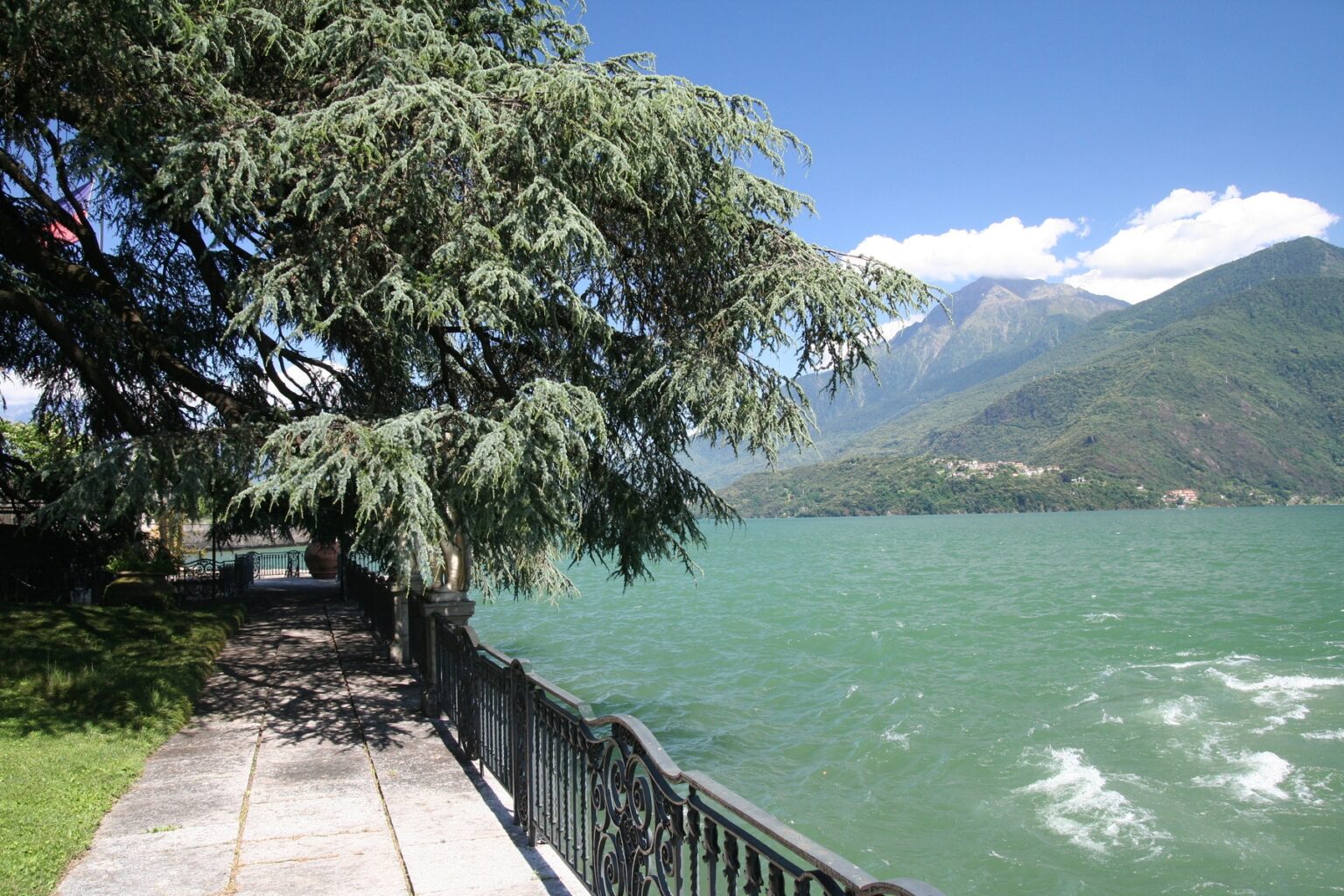 Scenic view of a lakeside promenade with a lush tree, overlooking a vibrant turquoise lake and mountains under a clear blue sky. | Only on SopranoVillas