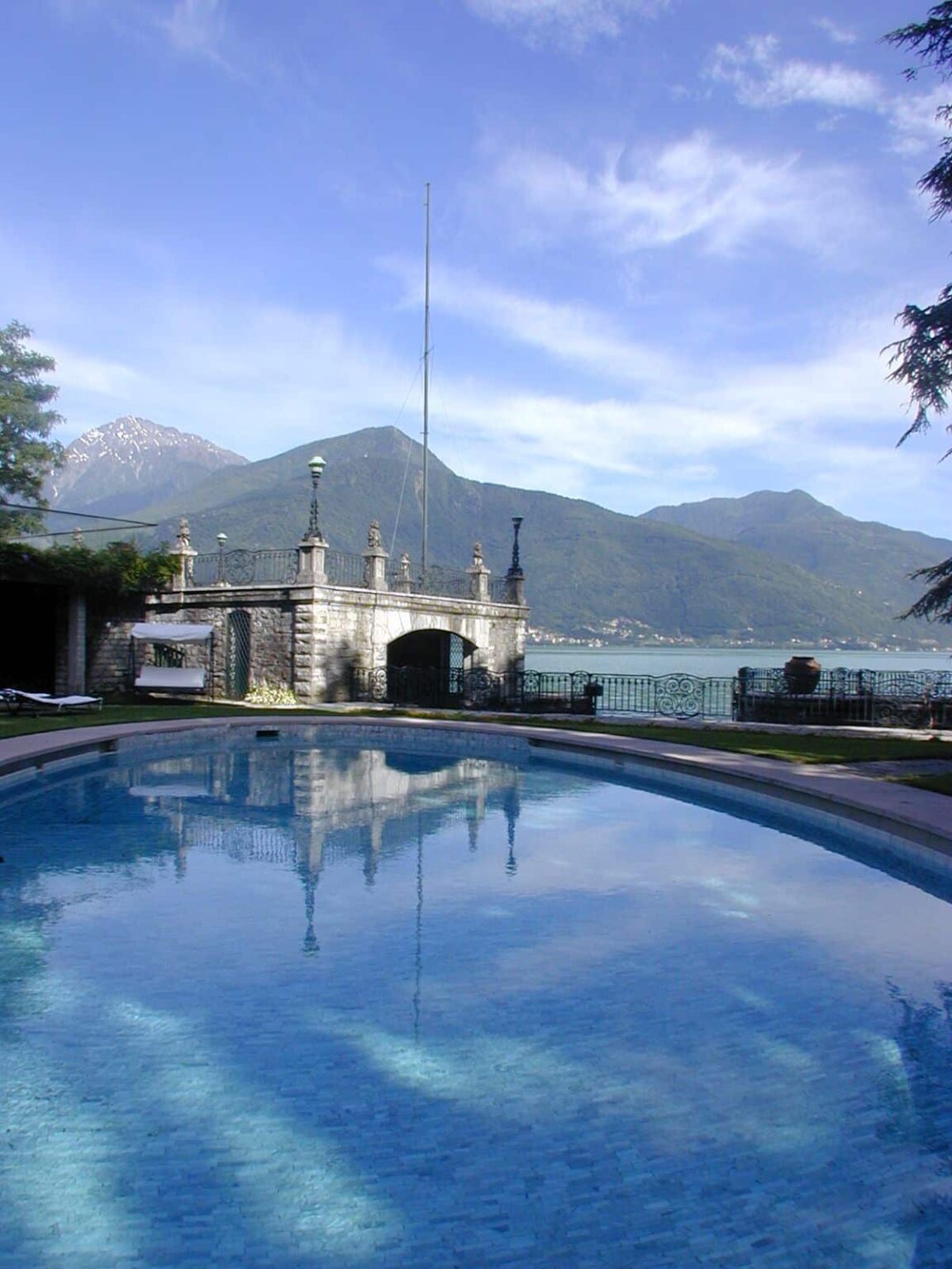 Scenic view of a swimming pool reflecting the surrounding mountains and lake, with a historic stone building in the background. | Only on SopranoVillas