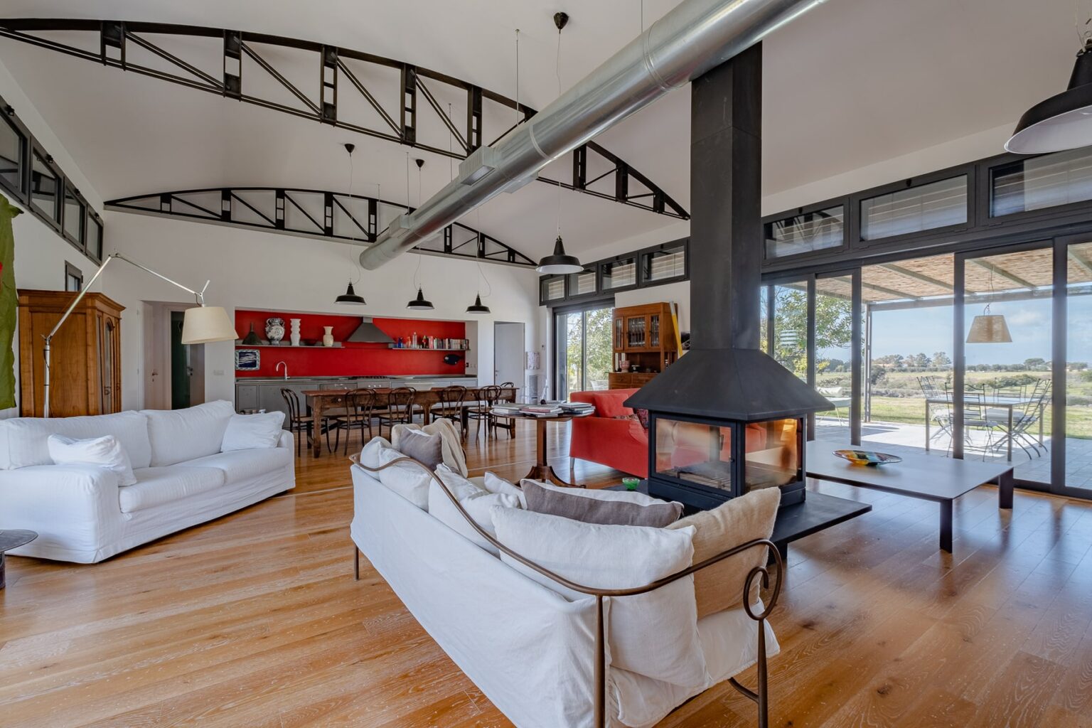 Modern living room with high ceilings, featuring a cozy fireplace, stylish white sofas, a wooden dining table, and large windows offering scenic views. The decor includes a red accent wall and contemporary lighting, creating a warm and inviting atmosphere. | Only on SopranoVillas