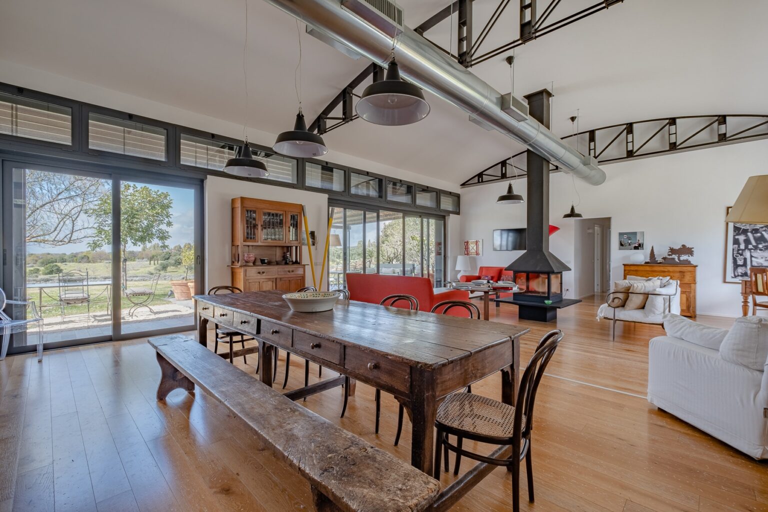 Spacious modern living area featuring a large wooden dining table, comfortable seating, and expansive windows that provide natural light and scenic views. The room showcases a blend of rustic and contemporary design elements with high ceilings and stylish decor. | Only on SopranoVillas