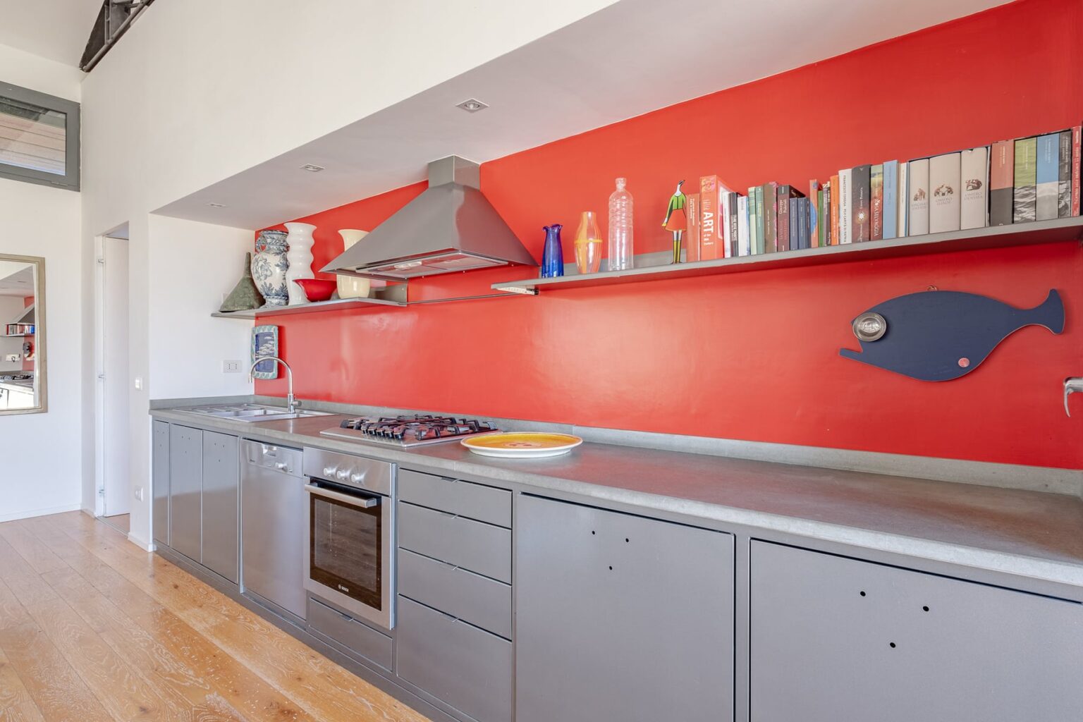 Modern kitchen featuring sleek gray cabinetry, a stainless steel oven, and a vibrant red accent wall with decorative shelves and a fish motif. The space is bright and inviting, highlighting contemporary design elements. | Only on SopranoVillas