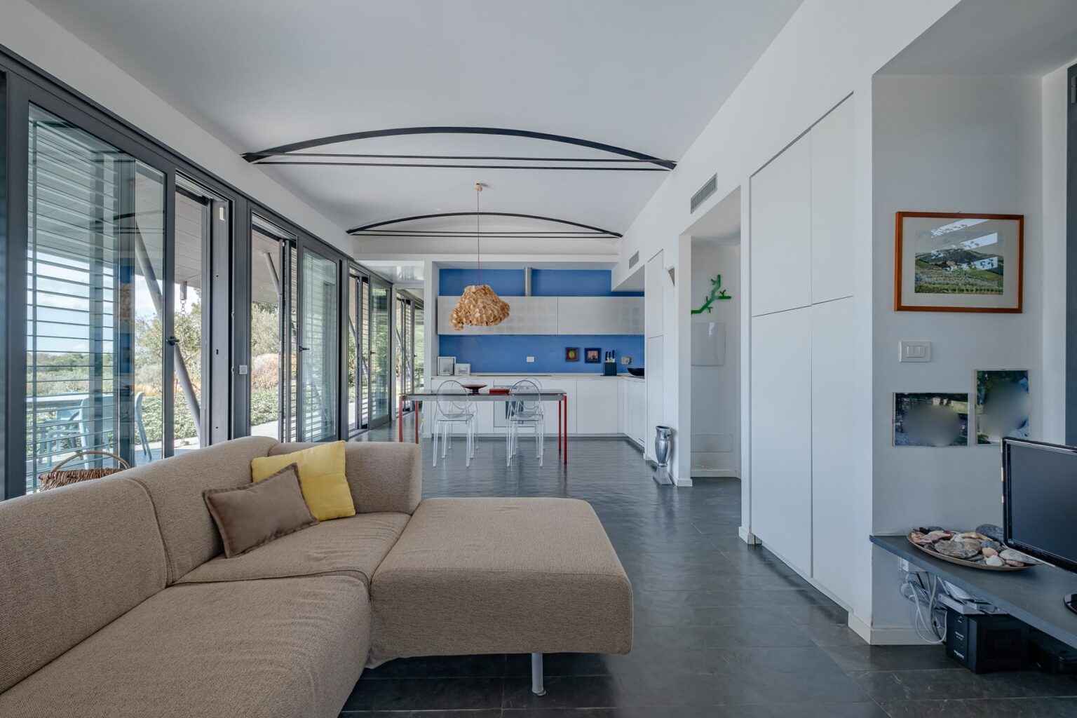 Modern living room featuring a beige sectional sofa with a yellow accent pillow, large windows allowing natural light, and a blue accent wall with kitchen space in the background. The design emphasizes an open layout and contemporary decor. | Only on SopranoVillas