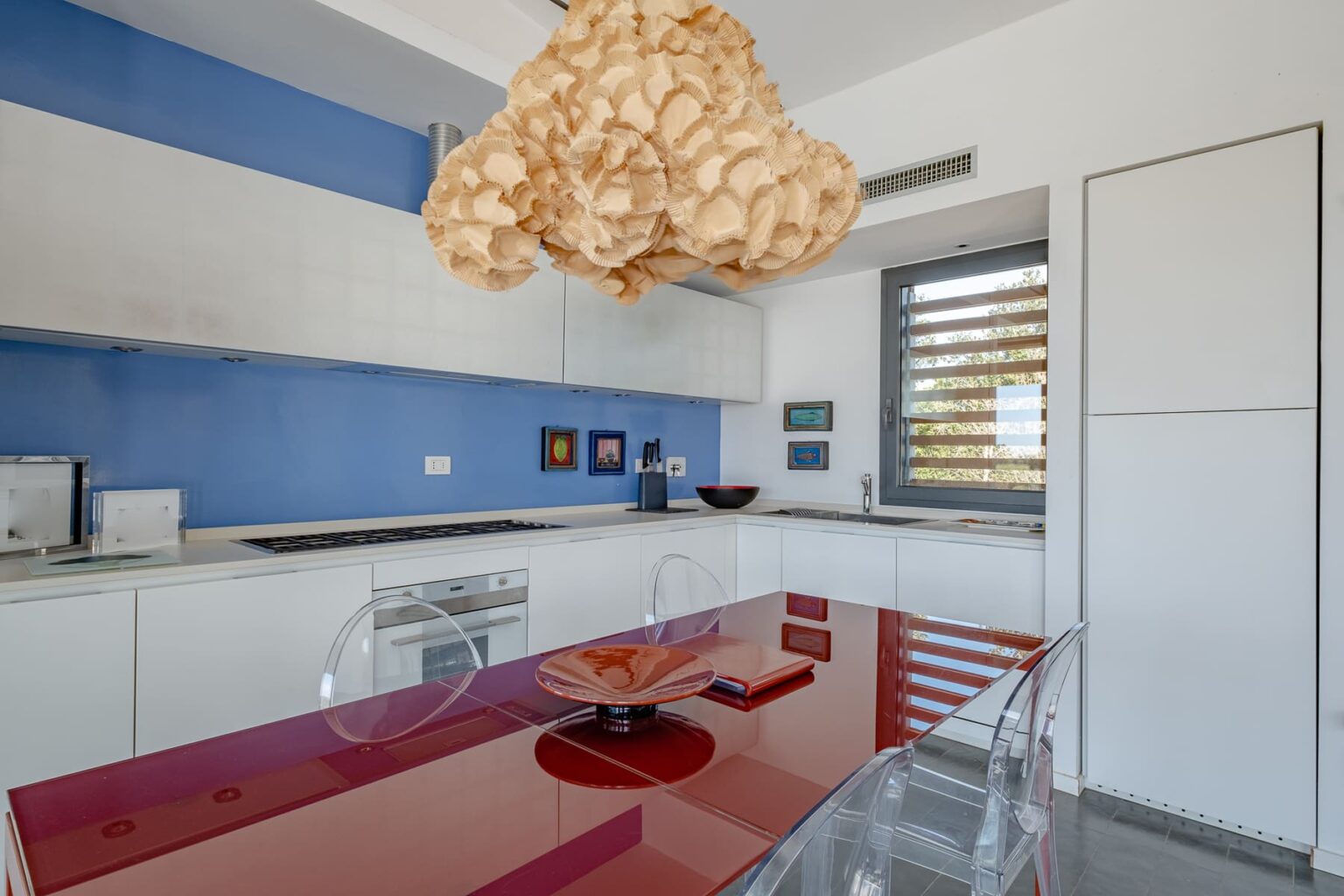 Modern kitchen interior featuring a blue accent wall, sleek white cabinetry, and a unique pendant light. A red glass dining table is set with minimalist decor, complemented by transparent chairs, creating a stylish and contemporary atmosphere. | Only on SopranoVillas