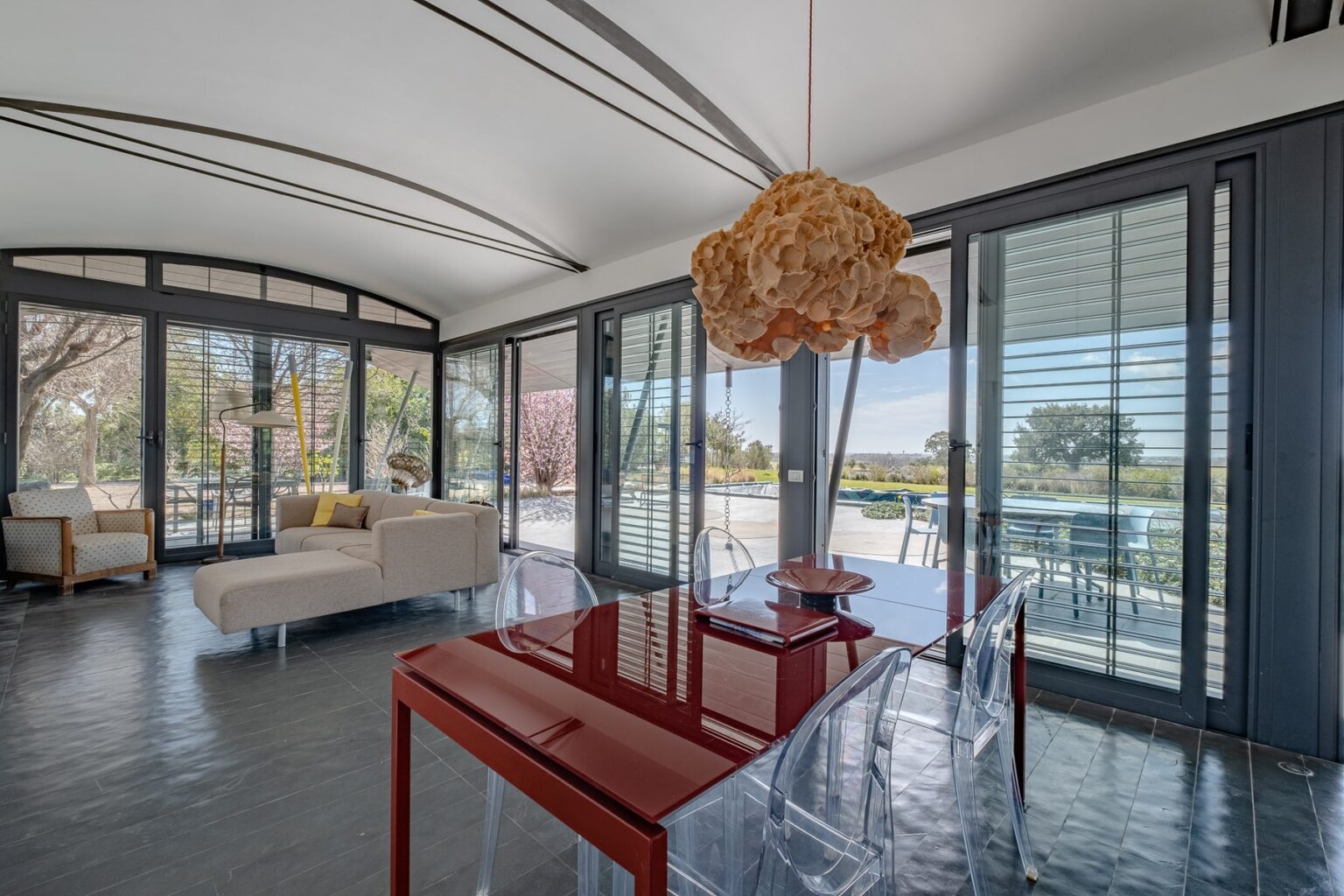 Modern sunlit living space featuring large glass windows, a stylish red dining table, and contemporary decor. The room is designed for comfort and elegance, showcasing a blend of indoor and outdoor views. | Only on SopranoVillas