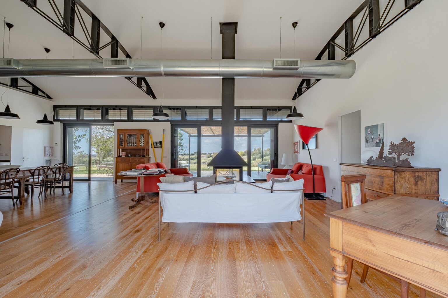 Spacious modern living room featuring high ceilings, large windows, and wooden flooring, furnished with a white sofa, red armchairs, and a central fireplace, creating a bright and inviting atmosphere. | Only on SopranoVillas