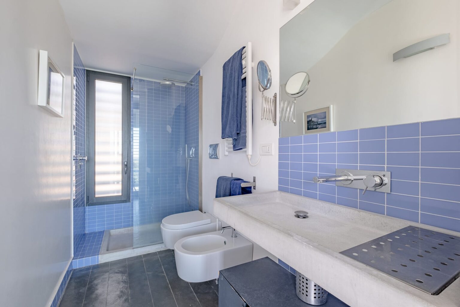 Modern bathroom featuring a sleek design with blue tiled walls, a spacious shower area, minimalist fixtures, and a stylish vanity with a large mirror. | Only on SopranoVillas