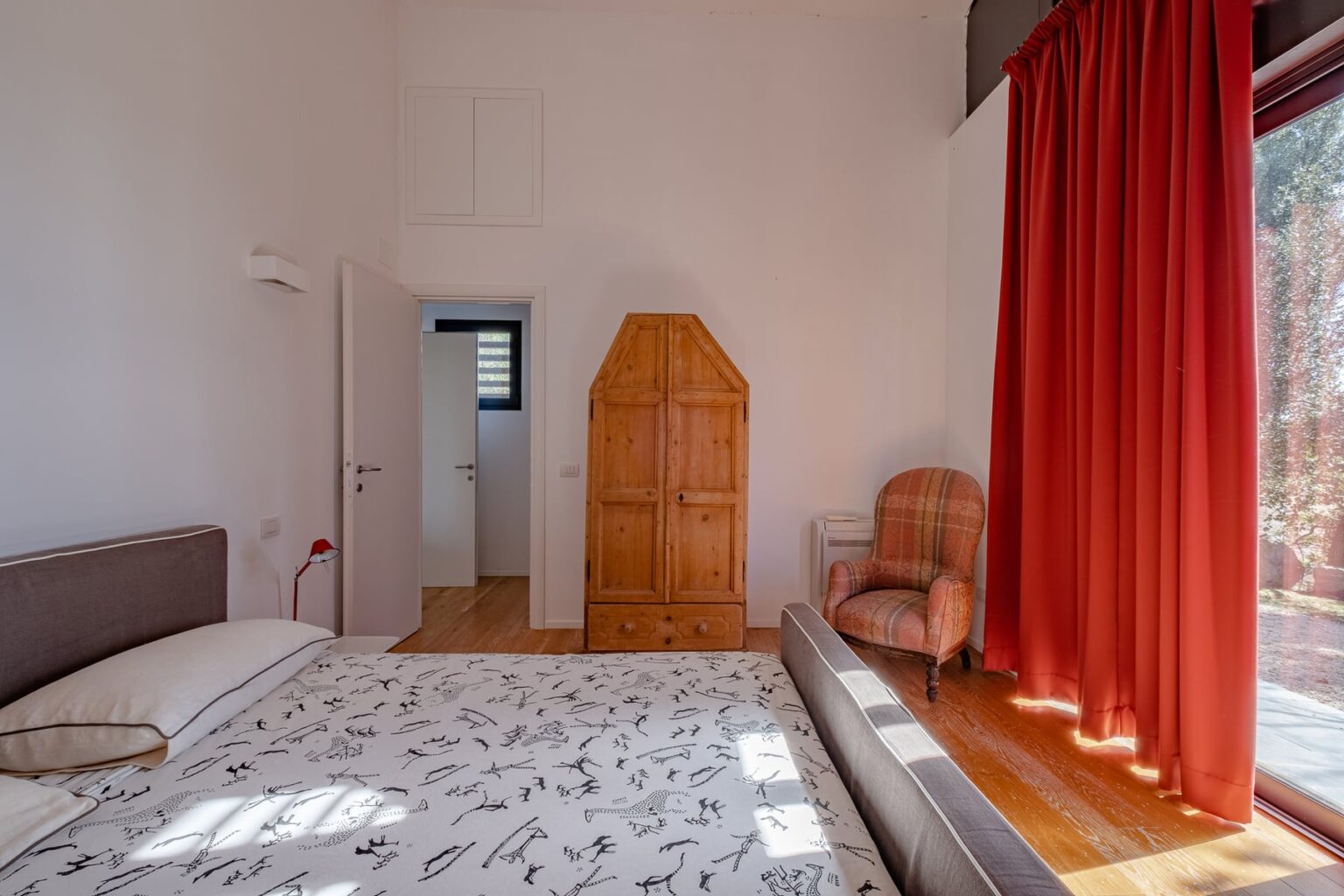 Bright and airy bedroom featuring a comfortable bed with patterned bedding, a wooden wardrobe, a cozy armchair, and large windows draped with red curtains, creating a warm and inviting atmosphere. | Only on SopranoVillas