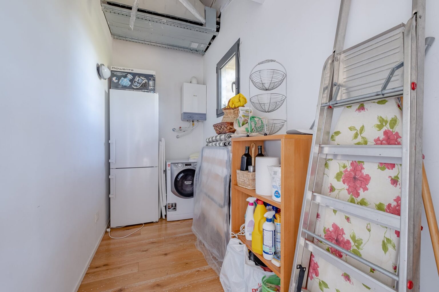 Laundry room with a white refrigerator, washing machine, and organized shelving displaying cleaning supplies. A folding ladder leans against the wall, and the space features wooden flooring and bright lighting. | Only on SopranoVillas