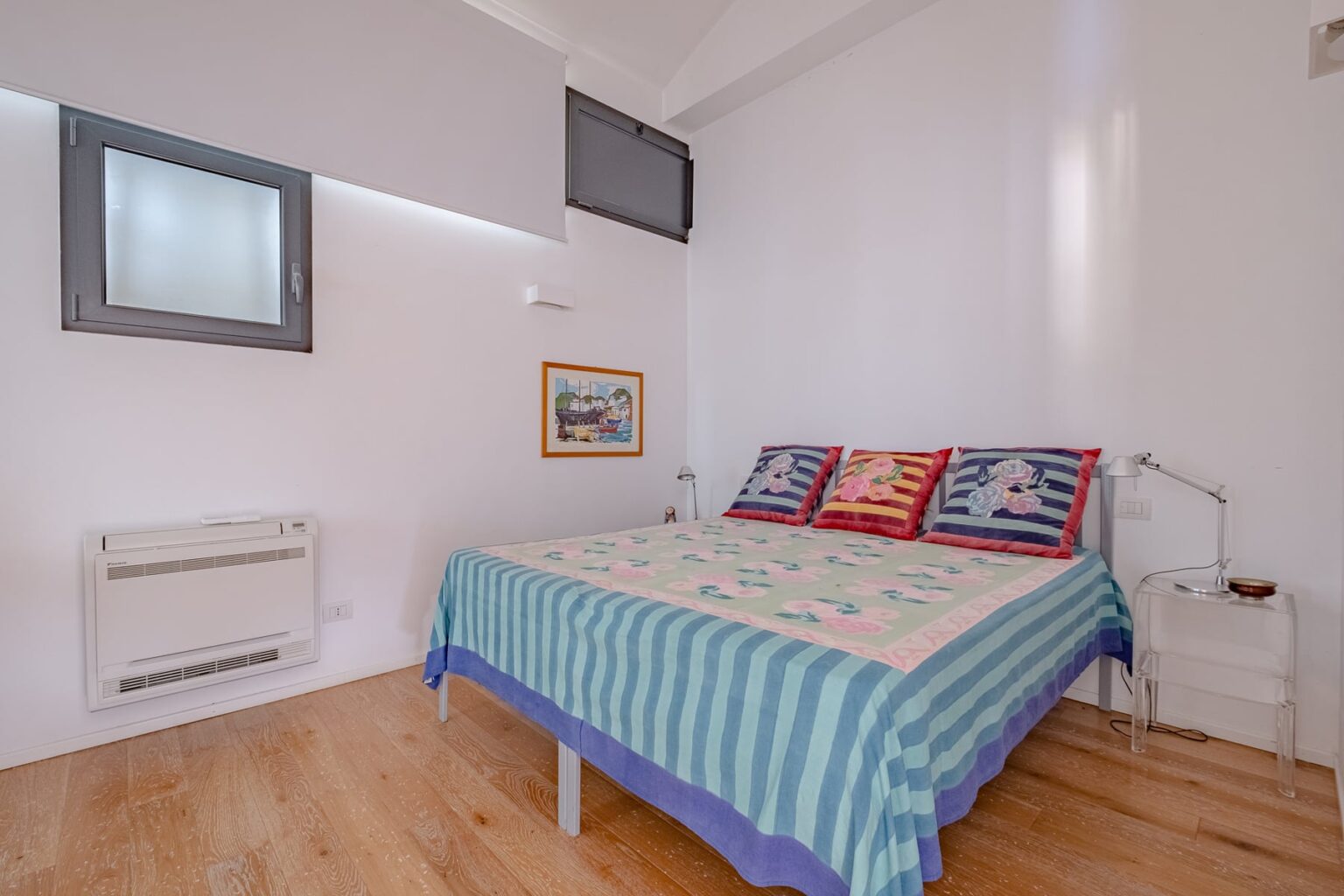 Cozy bedroom featuring a double bed with a colorful quilt, wooden flooring, and natural light from a window, complemented by modern decor and a small bedside table. | Only on SopranoVillas