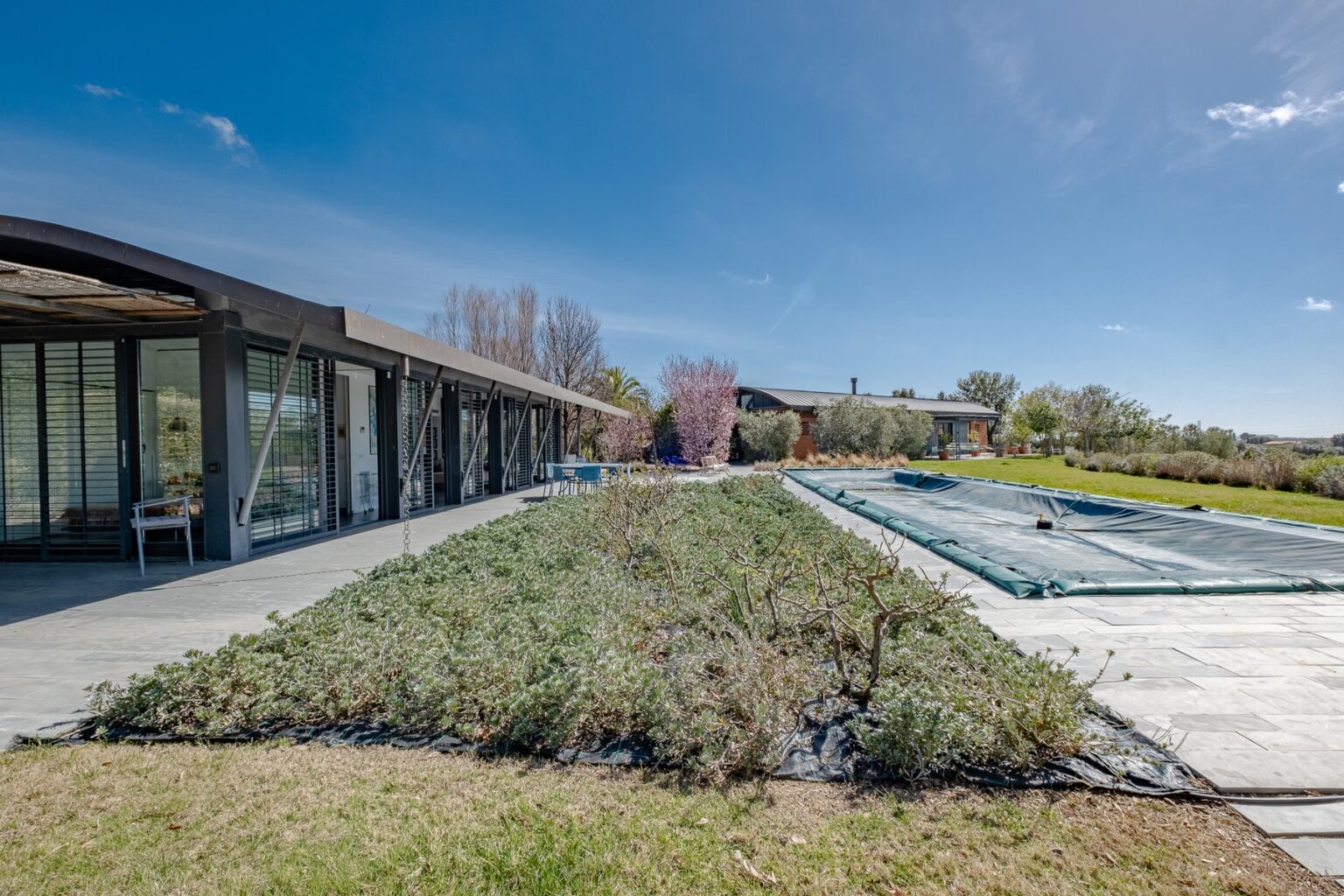 Modern architectural home with expansive glass walls, featuring a landscaped garden and a covered swimming pool under a clear blue sky. | Only on SopranoVillas