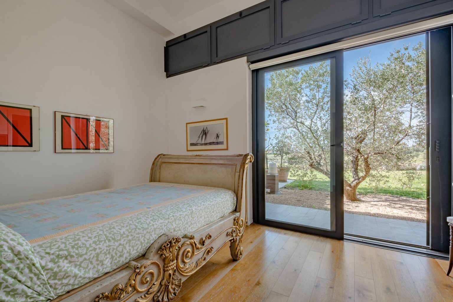 Bright and airy bedroom featuring a vintage sleigh bed, hardwood flooring, and large sliding glass doors that open to an outdoor view of olive trees. The room is adorned with framed artwork, enhancing its modern yet rustic charm. | Only on SopranoVillas