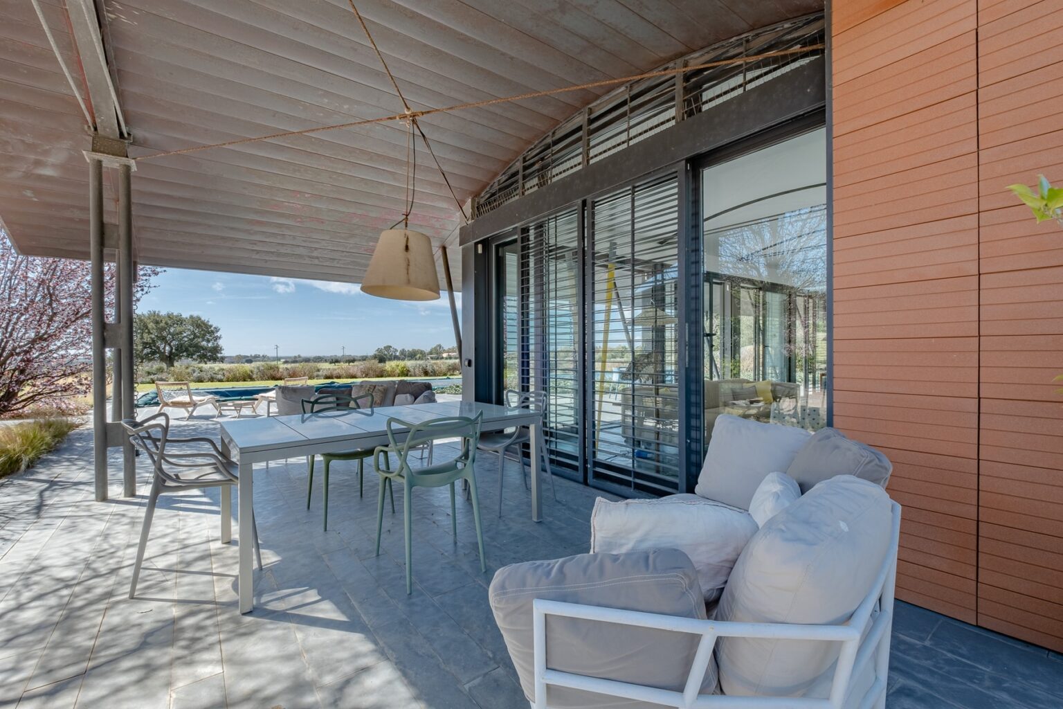 Modern outdoor patio with a dining table and chairs, featuring a cozy seating area, large windows, and scenic views of a landscaped garden. | Only on SopranoVillas