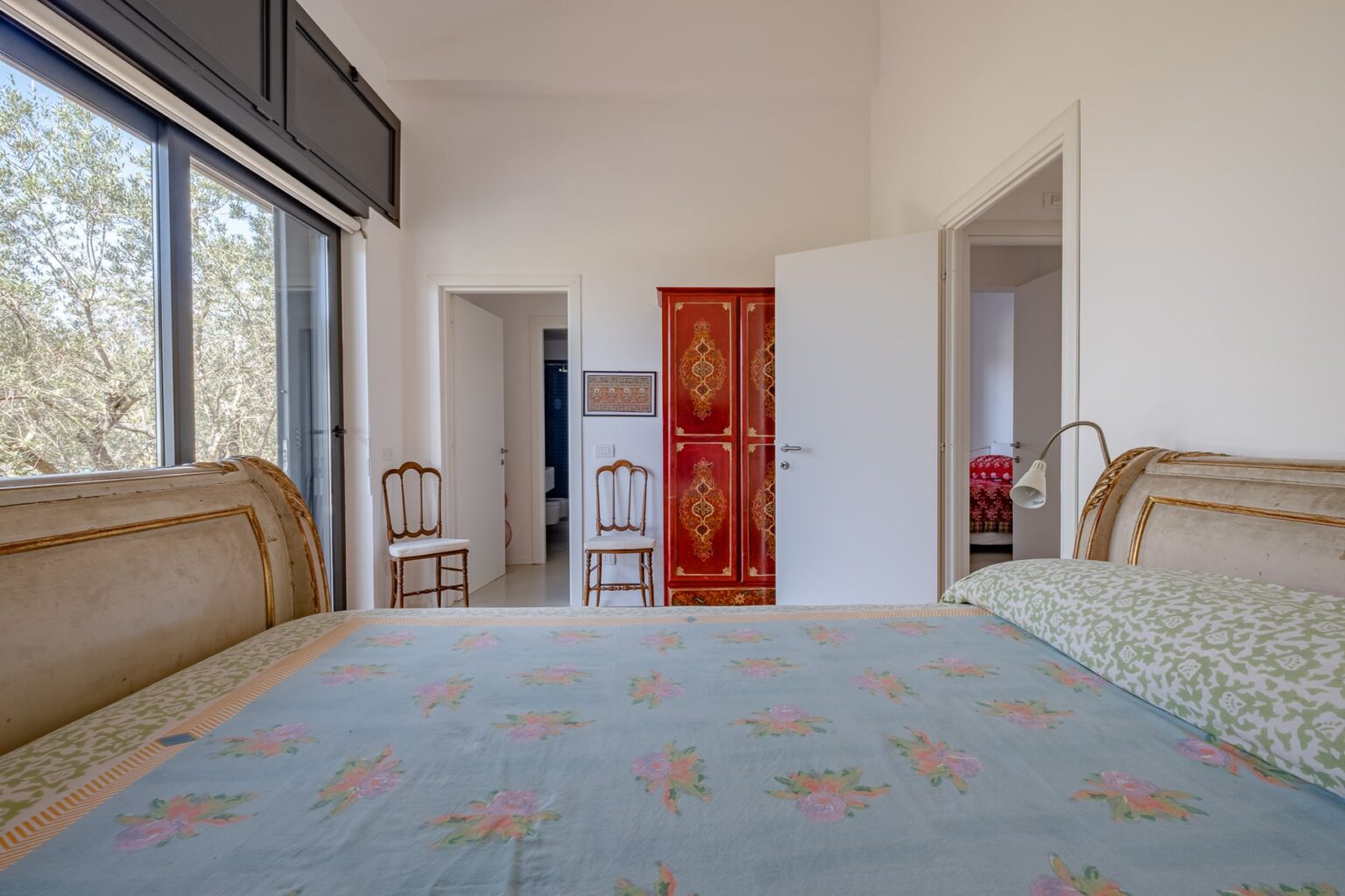 Cozy bedroom interior featuring a double bed with floral bedding, a vintage-style headboard, and a bright atmosphere. The room includes large windows with views of greenery, a stylish red wardrobe, and wooden chairs, creating a welcoming and serene environment. | Only on SopranoVillas