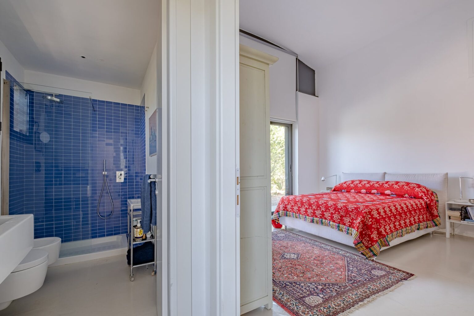 Modern bathroom with blue tiled shower and elegant bedroom featuring a red patterned bedspread and large windows for natural light. | Only on SopranoVillas