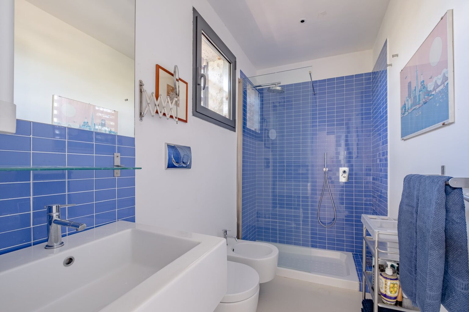 Modern bathroom featuring blue tiled walls, a sleek white sink, and a glass-enclosed shower. The space includes a bidet, a small shelving unit with toiletries, and decorative wall art, creating a stylish and functional design. | Only on SopranoVillas