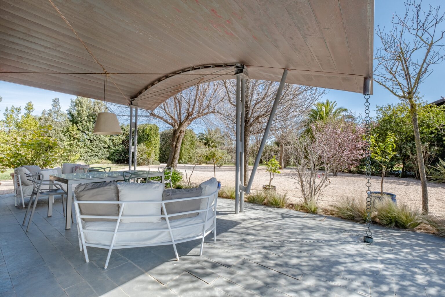 Outdoor patio area featuring modern white furniture, a glass dining table, and a hanging lamp, surrounded by lush greenery and flowering trees, ideal for relaxation and dining al fresco. | Only on SopranoVillas