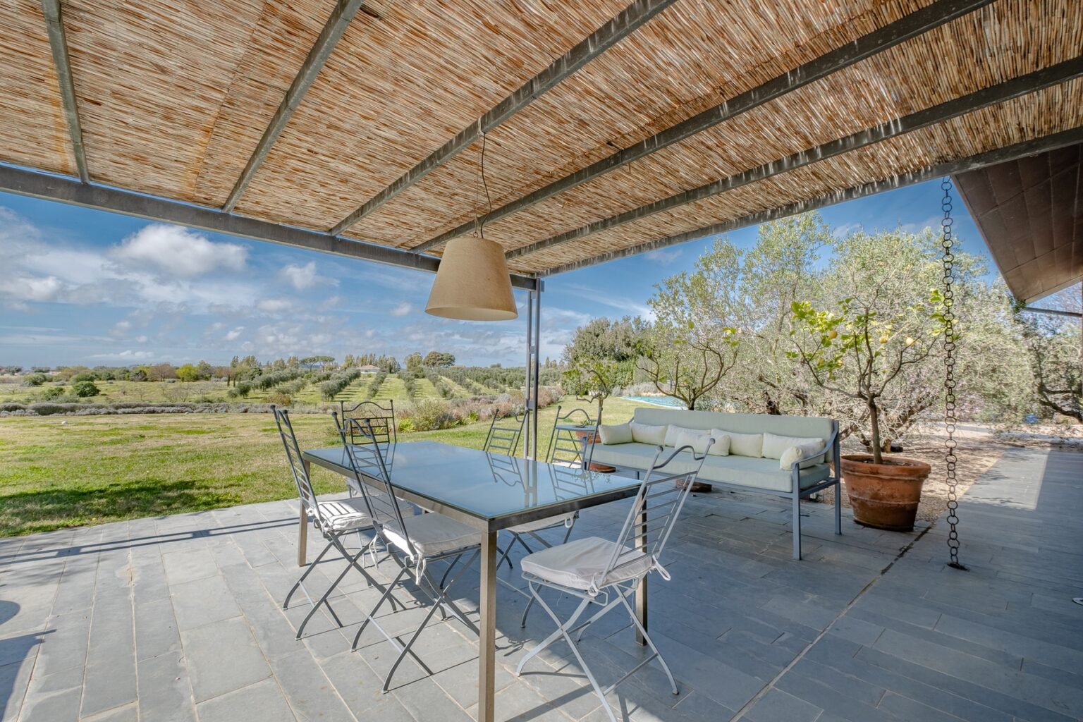 Modern outdoor dining area with a glass table and metal chairs under a thatched roof, overlooking a scenic landscape with greenery and blue skies. Ideal for alfresco dining and relaxation. | Only on SopranoVillas