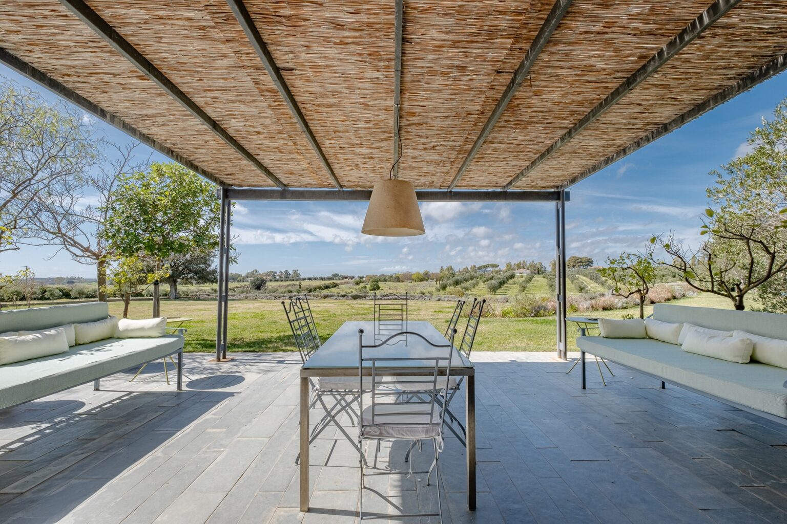 Outdoor dining area with a large table and metal chairs under a thatched roof, surrounded by greenery and scenic views of a landscape. Ideal for gatherings and relaxation in a tranquil setting. | Only on SopranoVillas