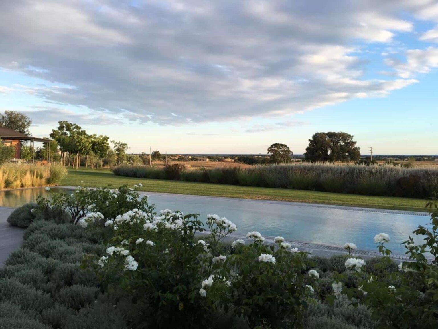 Scenic view of a tranquil landscape featuring a swimming pool surrounded by lush greenery and flowering shrubs, with a clear sky and distant hills in the background. Ideal for relaxation and outdoor living. | Only on SopranoVillas