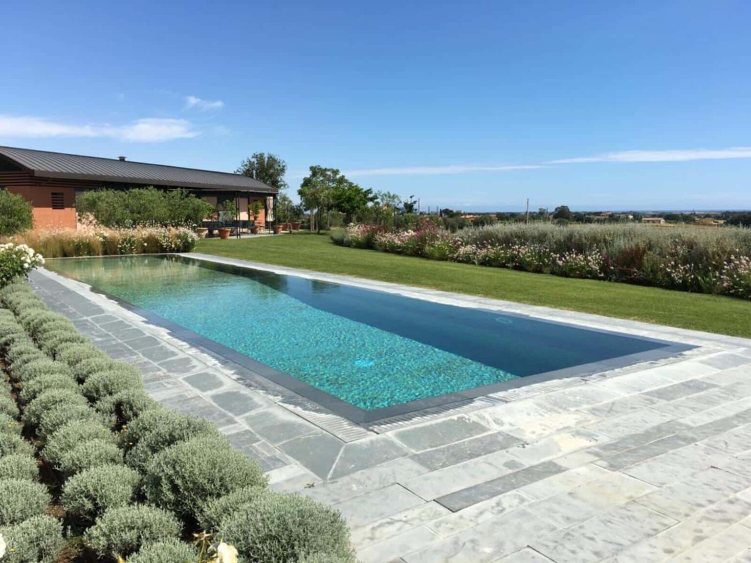 Modern outdoor swimming pool surrounded by lush greenery and landscaped gardens, with a clear blue sky in the background. Ideal for luxury home designs and outdoor living inspiration. | Only on SopranoVillas