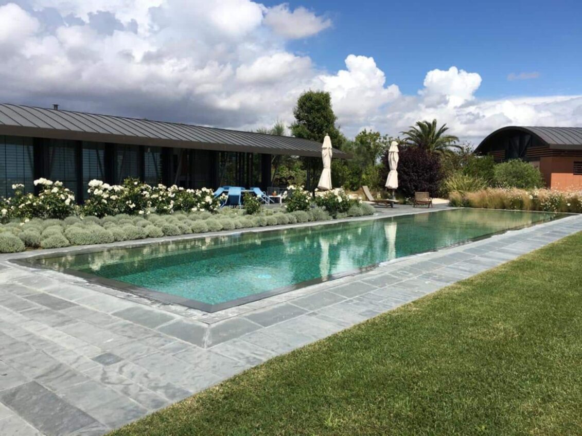Modern outdoor swimming pool surrounded by lush greenery and landscaped gardens, with a sleek stone patio and a contemporary home in the background under a partly cloudy sky. | Only on SopranoVillas