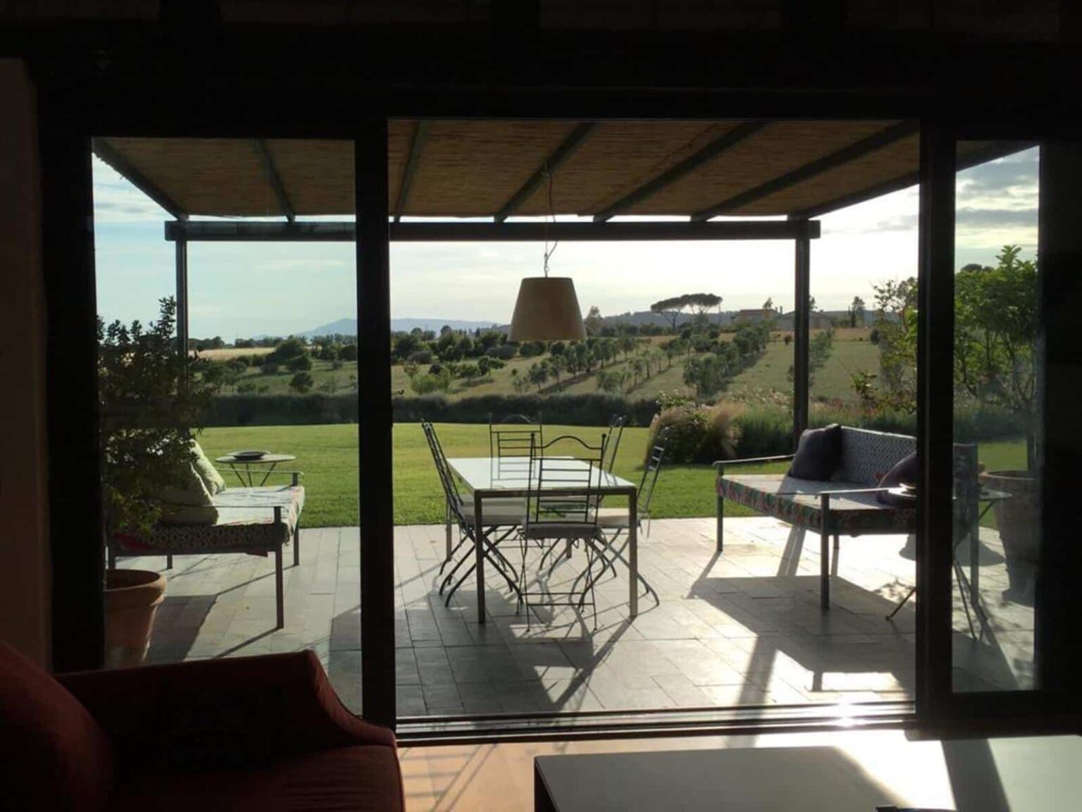 View from a modern living room showcasing a patio with outdoor furniture, overlooking a lush green landscape and distant hills under a clear sky. | Only on SopranoVillas