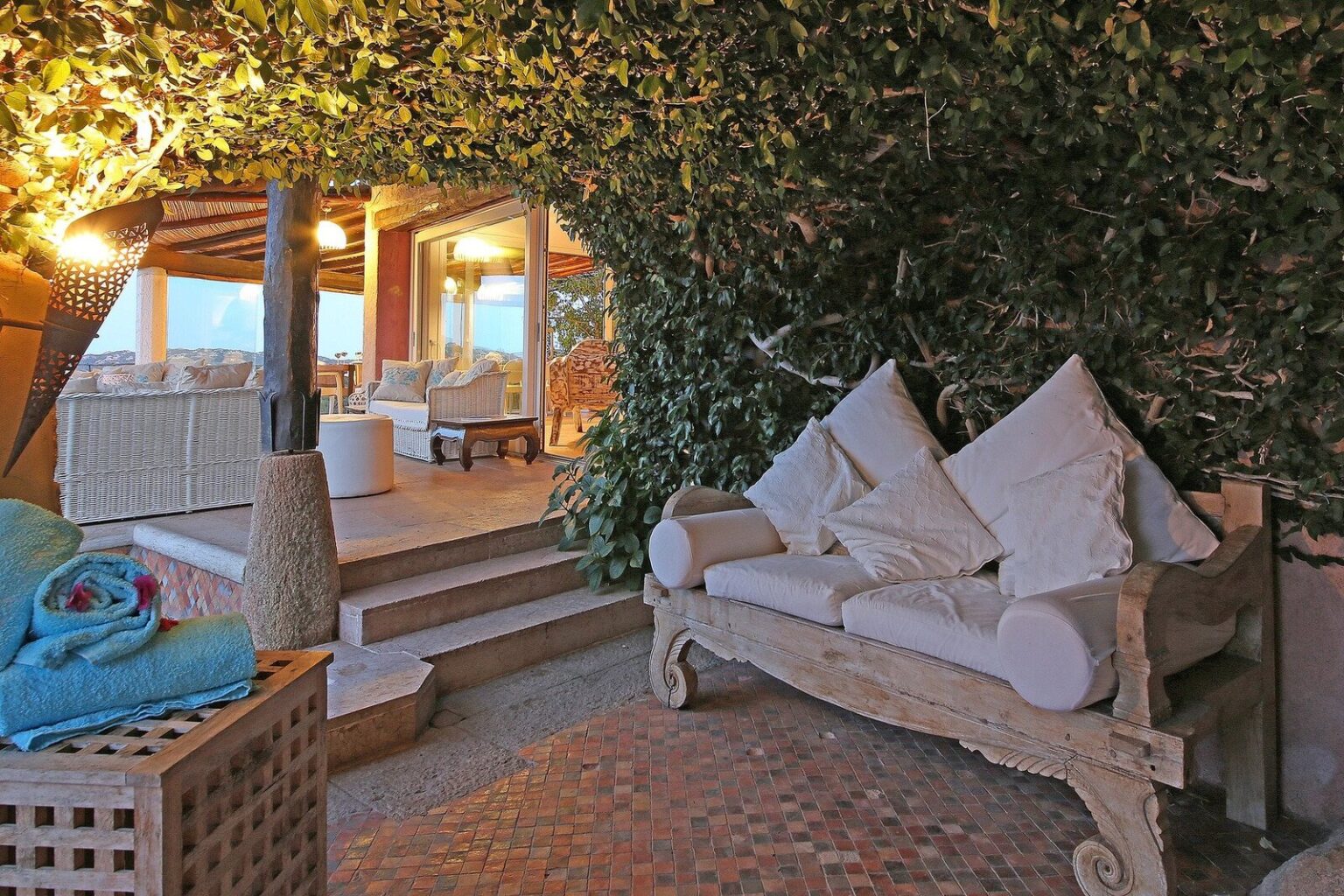 Cozy outdoor seating area featuring a wooden sofa with white cushions, surrounded by lush greenery, leading to a stylish indoor space with warm lighting. Ideal for relaxation and enjoying nature. | Only on SopranoVillas