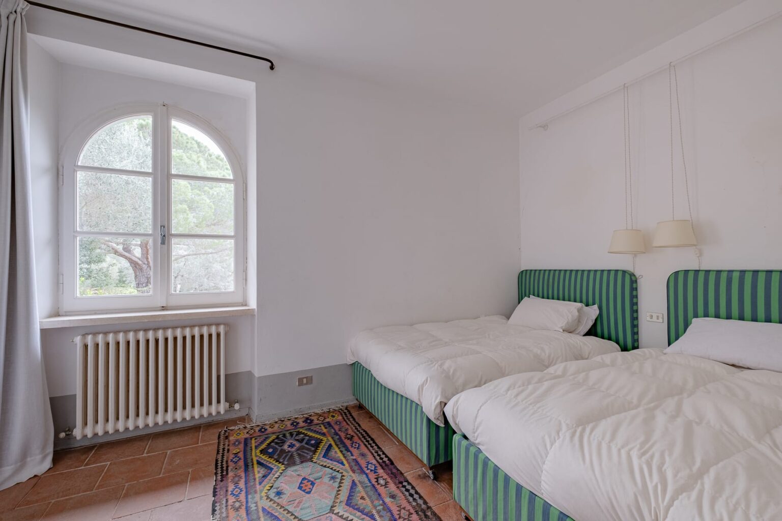 Cozy bedroom featuring two green upholstered twin beds, a large arched window with natural light, a patterned area rug, and soft white bedding, creating a serene and inviting atmosphere. | Only on SopranoVillas