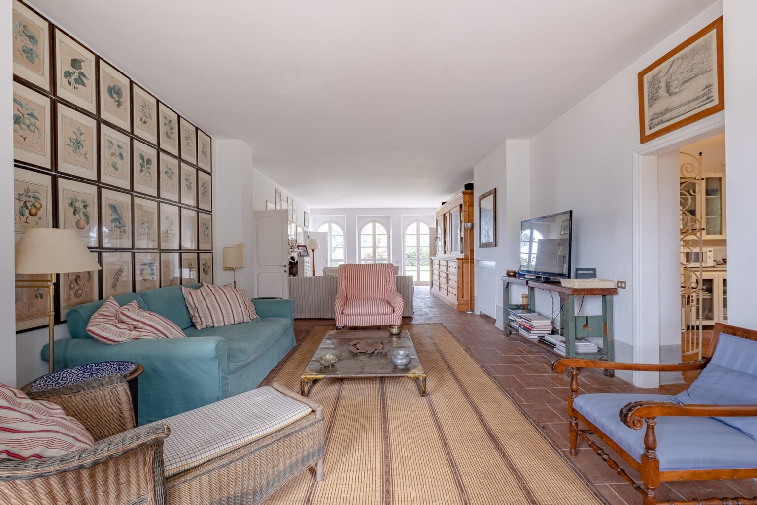 Spacious living room featuring comfortable seating with blue and striped chairs, a coffee table, and a large wall of framed artwork. Natural light fills the space, creating a warm and inviting atmosphere. | Only on SopranoVillas