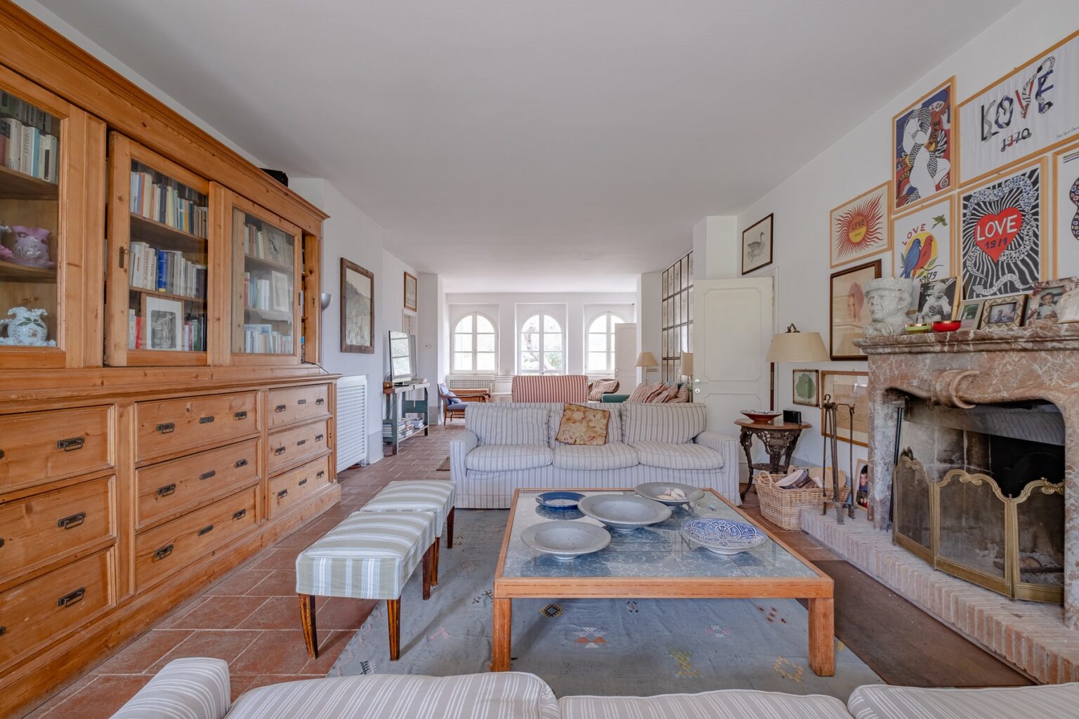 Spacious and inviting living room featuring a wooden bookshelf, cozy seating area, and a stylish coffee table with decorative items. Natural light fills the room through large windows, enhancing the warm decor and artwork on the walls. | Only on SopranoVillas