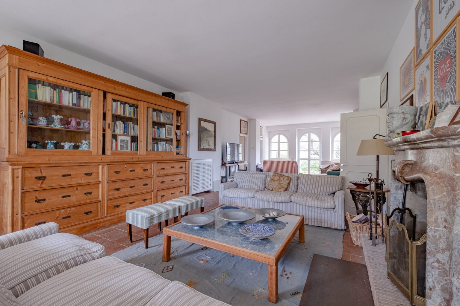 Spacious living room featuring a wooden display cabinet, comfortable sofas, a coffee table, and a cozy fireplace, with large windows providing ample natural light. | Only on SopranoVillas