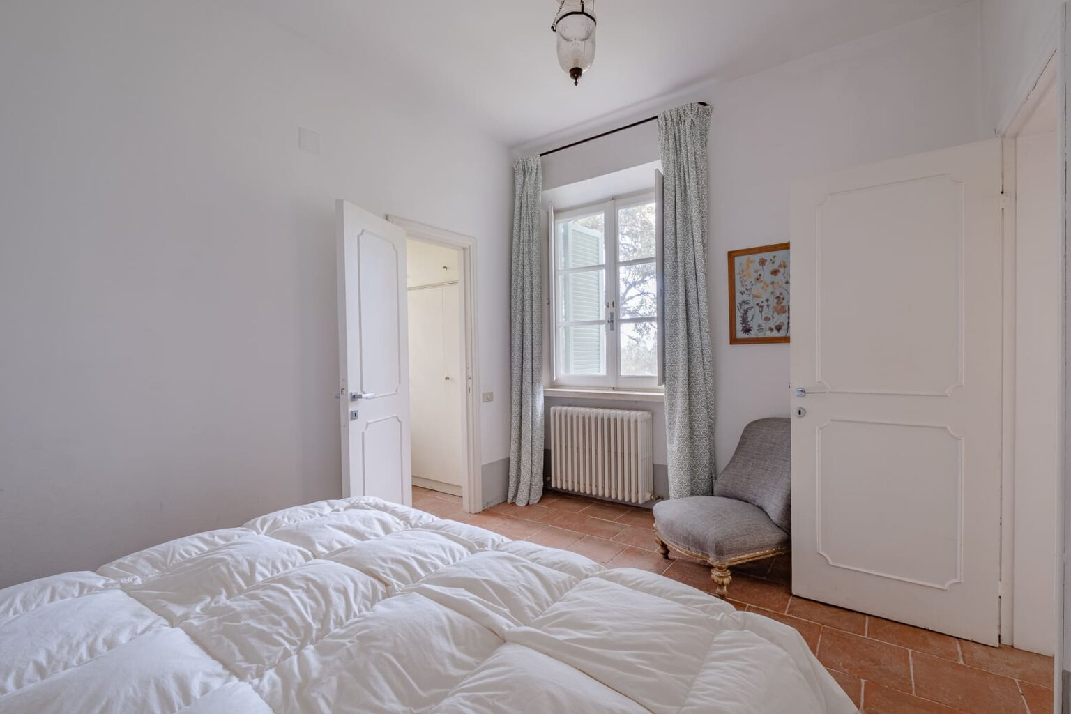 Bright and airy bedroom featuring a cozy bed with a white comforter, a window with light curtains, a comfortable chair, and two doors leading to different areas. The room has a warm, inviting atmosphere with neutral-colored walls and natural light. | Only on SopranoVillas