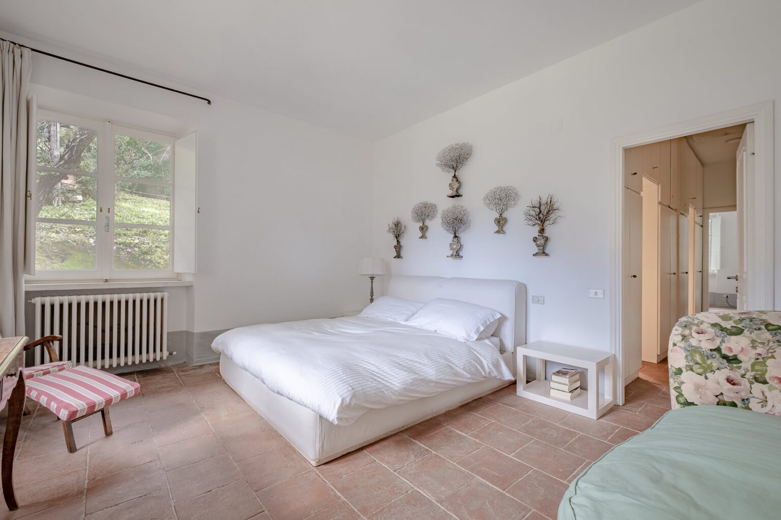 Bright and airy bedroom featuring a large bed, decorative wall art, a small desk with a chair, and a window allowing natural light to fill the space. The room has a minimalist design with neutral colors and warm wooden accents. | Only on SopranoVillas