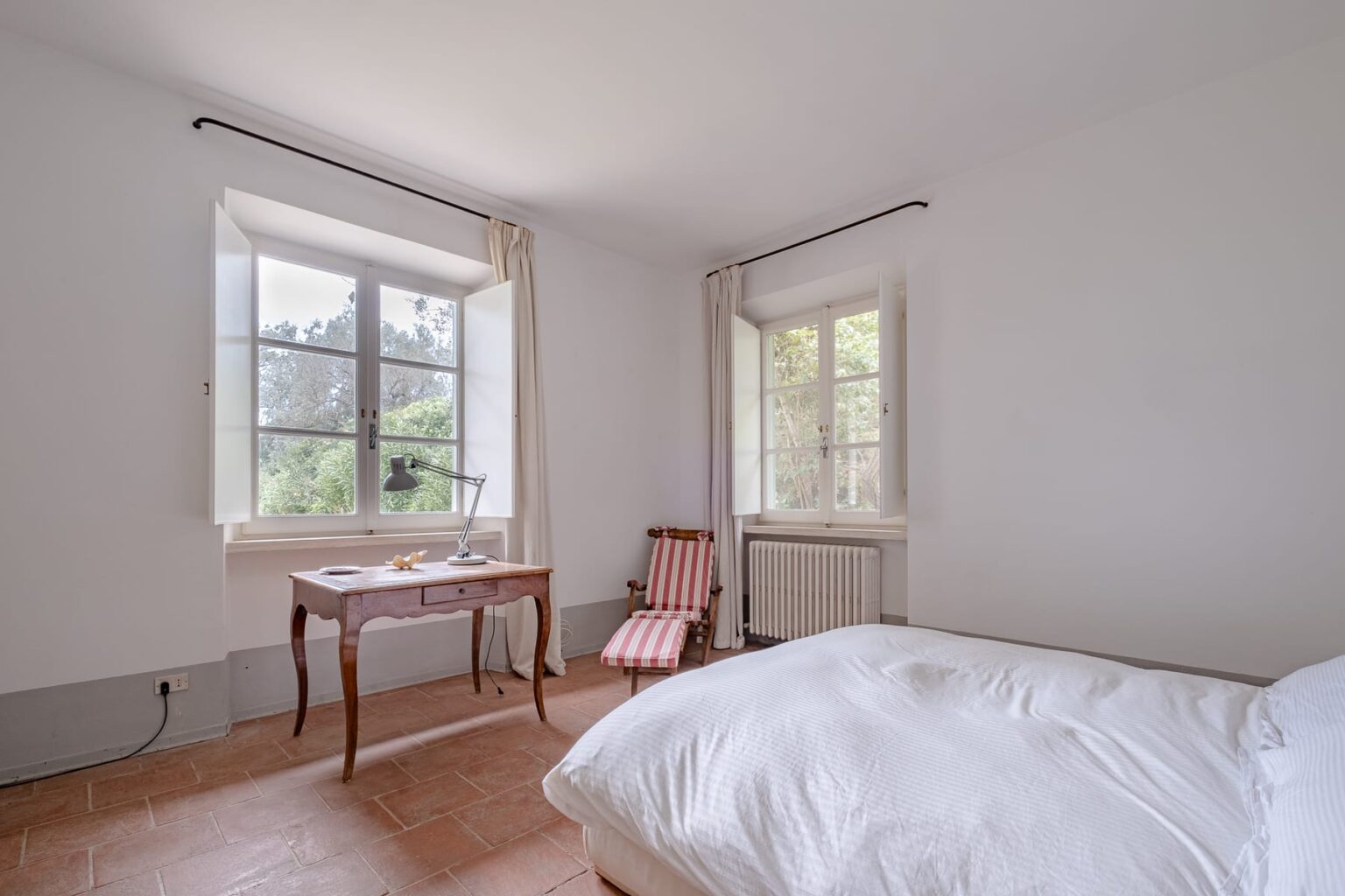 Bright and airy bedroom featuring a large window with natural light, a cozy bed with white bedding, a wooden desk, and a striped armchair, set against a minimalist white wall. | Only on SopranoVillas