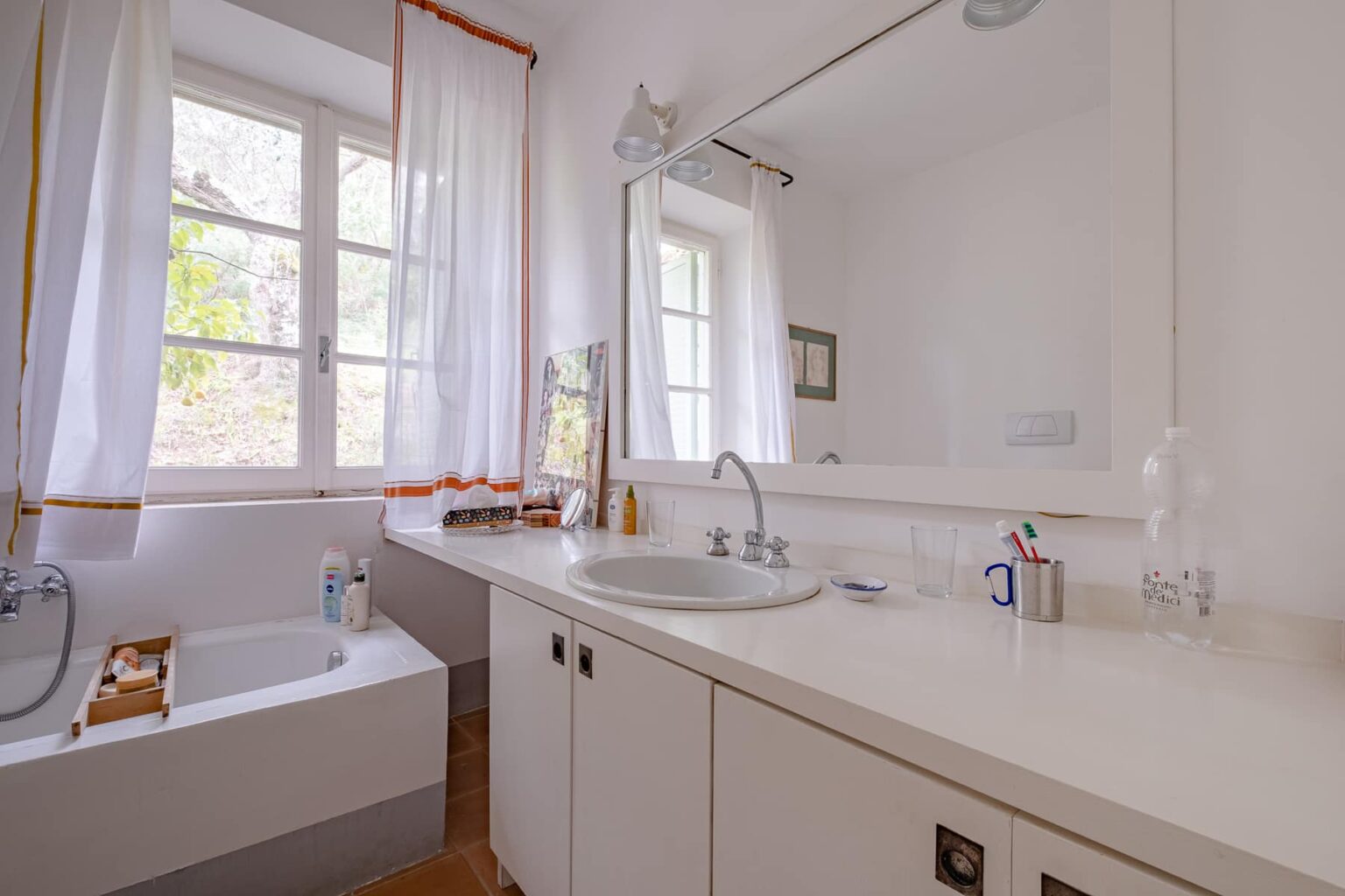 Bright and modern bathroom featuring a large mirror, white cabinetry, a bathtub, and natural light streaming through the window, creating an inviting atmosphere. | Only on SopranoVillas