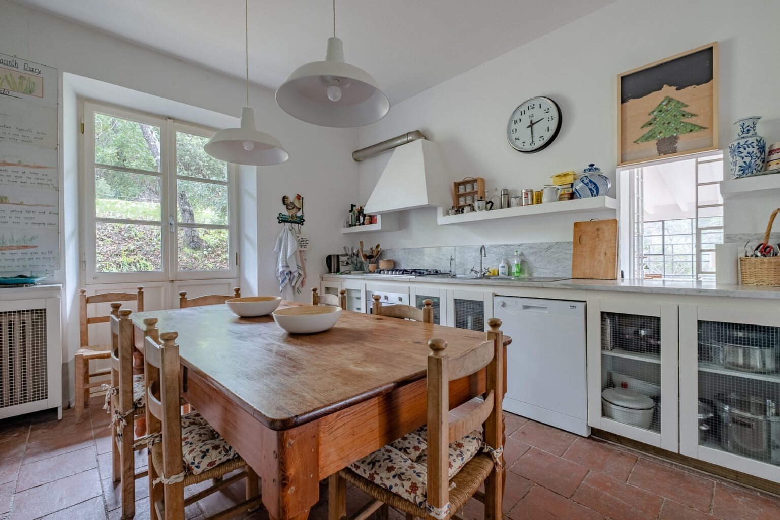 Bright and airy kitchen featuring a wooden dining table surrounded by chairs, white cabinetry, and modern appliances. Natural light streams through large windows, enhancing the inviting atmosphere. Decorative elements include a clock and wall art, adding charm to the space. | Only on SopranoVillas