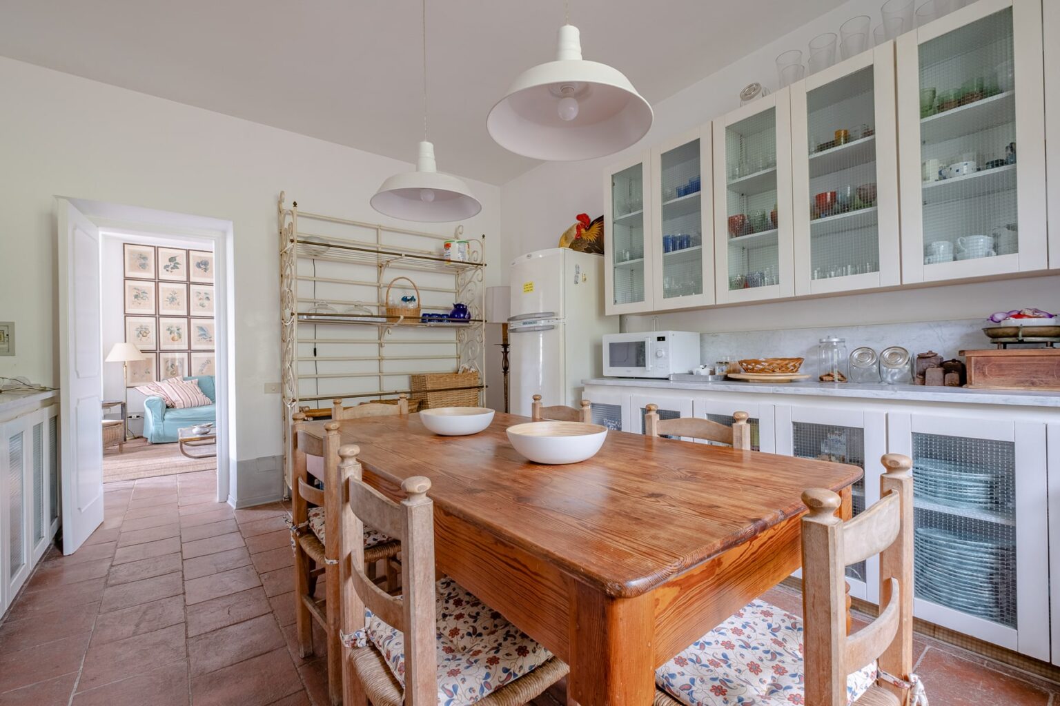 Bright and airy kitchen featuring a wooden dining table with bowls, white cabinetry, and modern appliances, complemented by pendant lighting and a view into a cozy living area. | Only on SopranoVillas