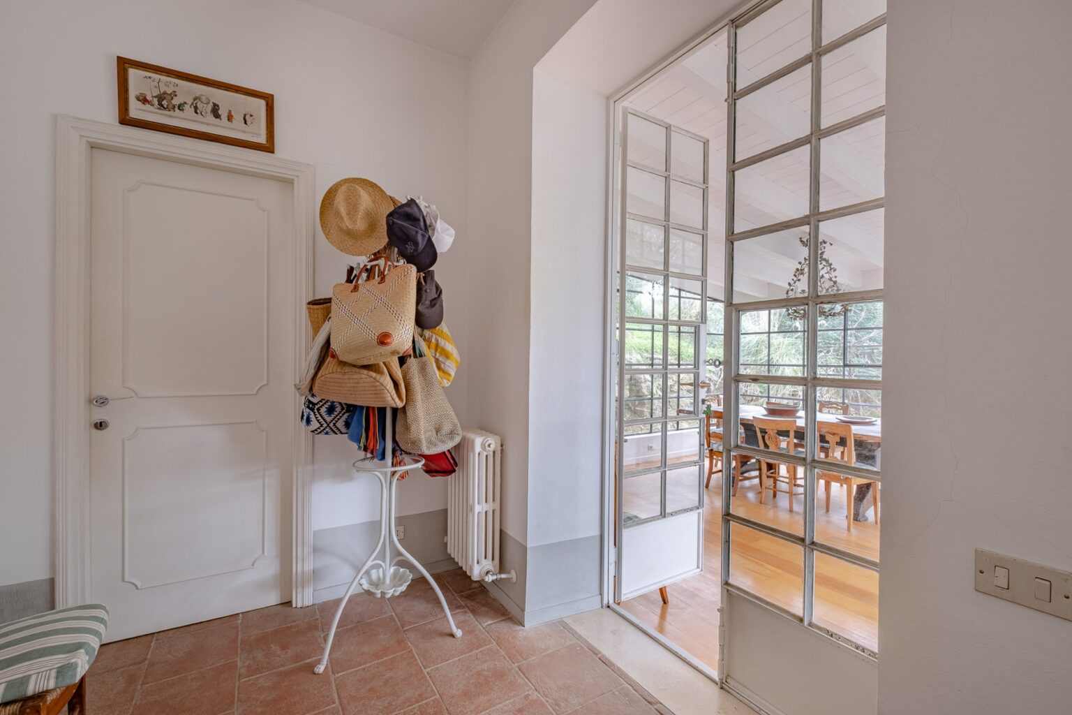 Entrance hallway featuring a coat rack adorned with various bags and hats, leading to a bright dining area visible through glass doors. The space has a warm, inviting atmosphere with tiled flooring and natural light. | Only on SopranoVillas