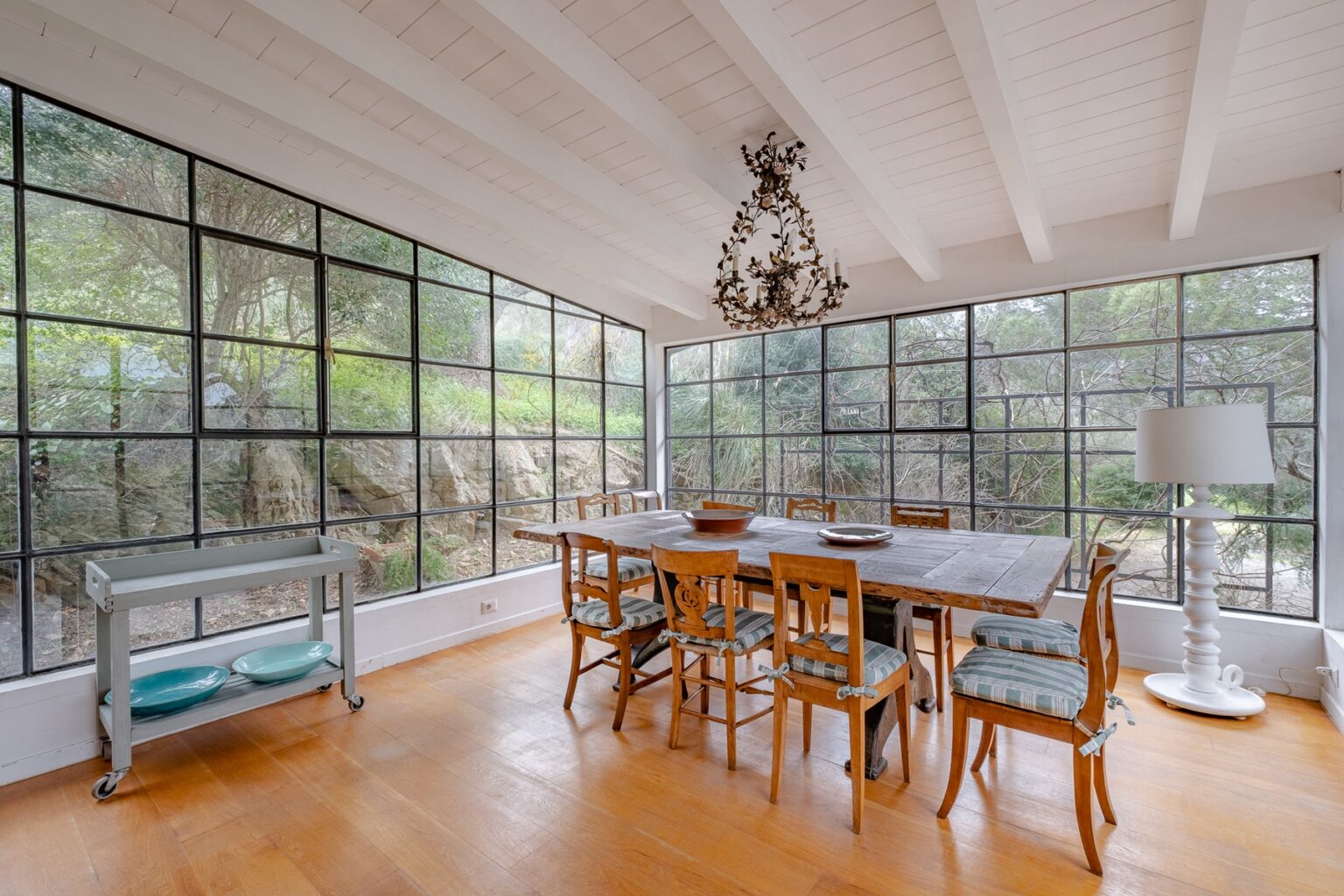 Bright and airy dining room featuring a large wooden table surrounded by vintage chairs, illuminated by a decorative chandelier. Floor-to-ceiling windows provide a view of the outdoor greenery, while a stylish lamp and a serving cart add functionality to the space. | Only on SopranoVillas