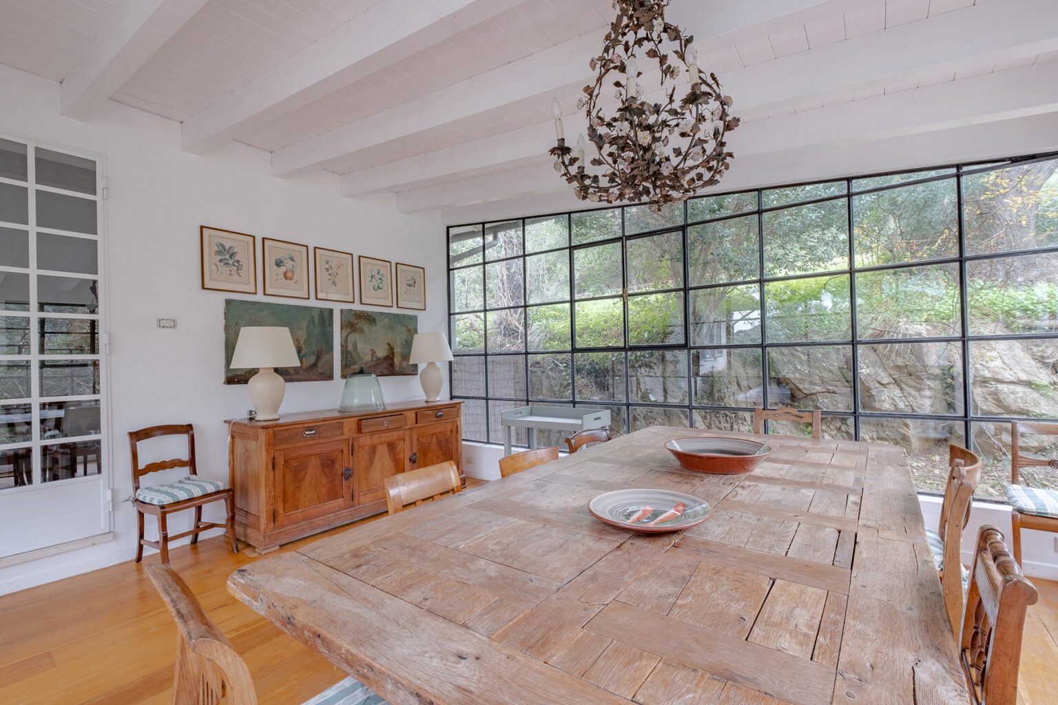 Spacious dining room featuring a large wooden table, vintage chairs, a decorative chandelier, and large glass windows that provide natural light and views of the outdoors. The decor includes framed artwork and a wooden sideboard. | Only on SopranoVillas