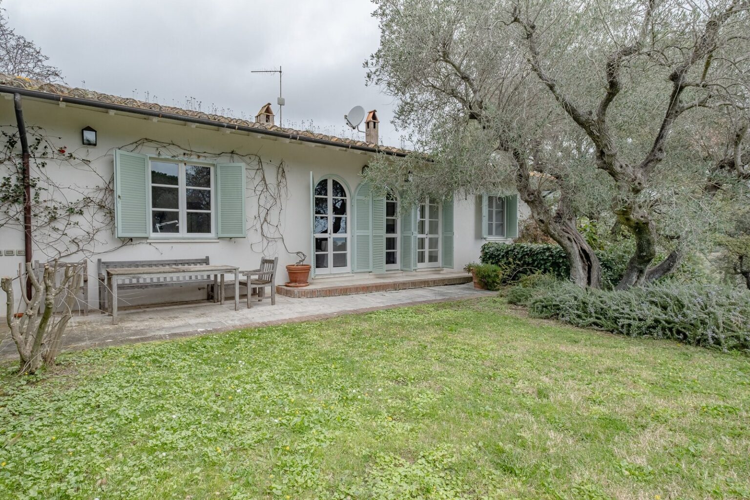 Charming countryside house with green shutters, a wooden bench, and a well-maintained garden, surrounded by olive trees and a cloudy sky. Ideal for relaxing outdoor gatherings. | Only on SopranoVillas