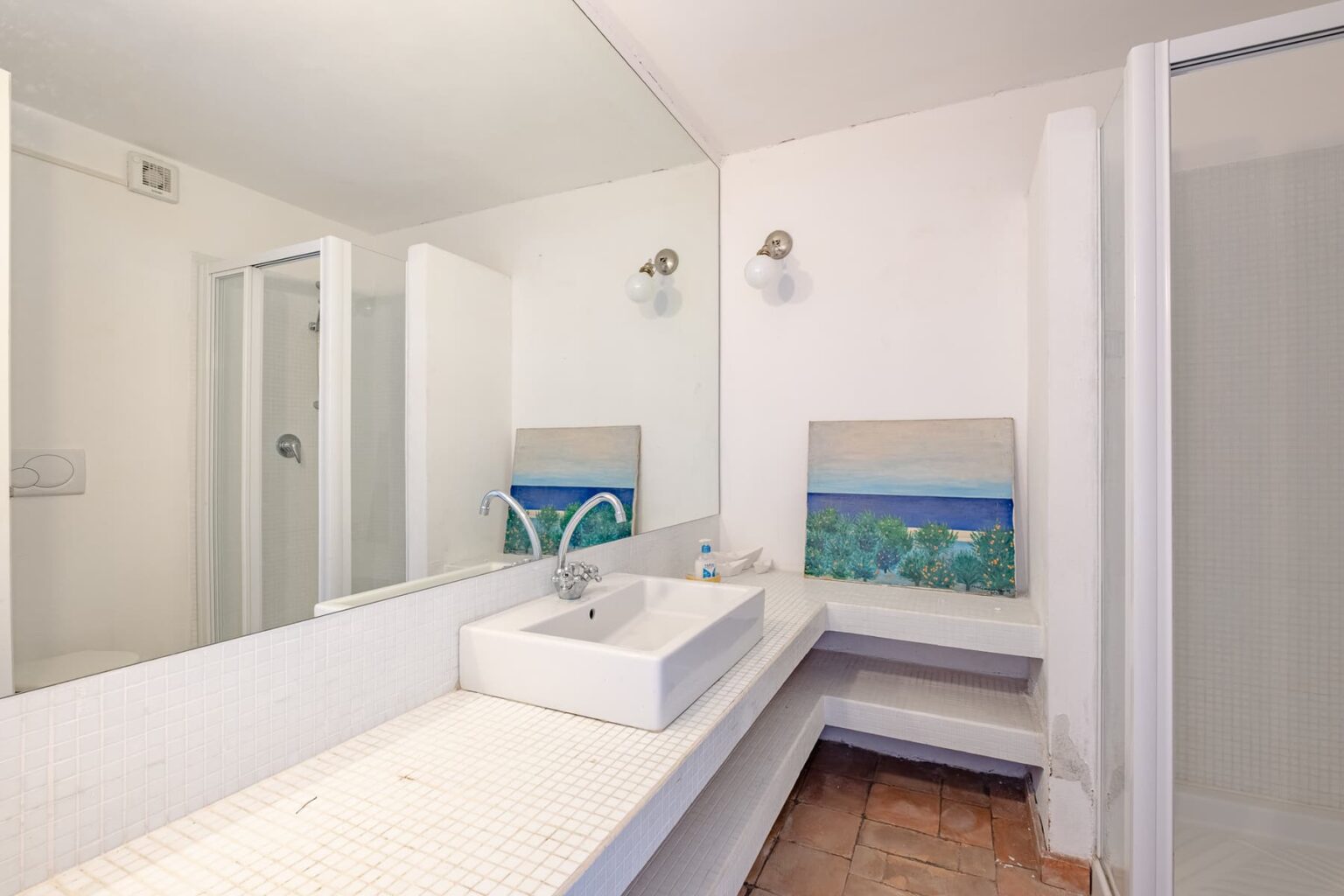 Modern bathroom interior featuring a minimalist design, white cabinetry, a rectangular sink, and a shower area, complemented by ocean-themed artwork and natural light. | Only on SopranoVillas