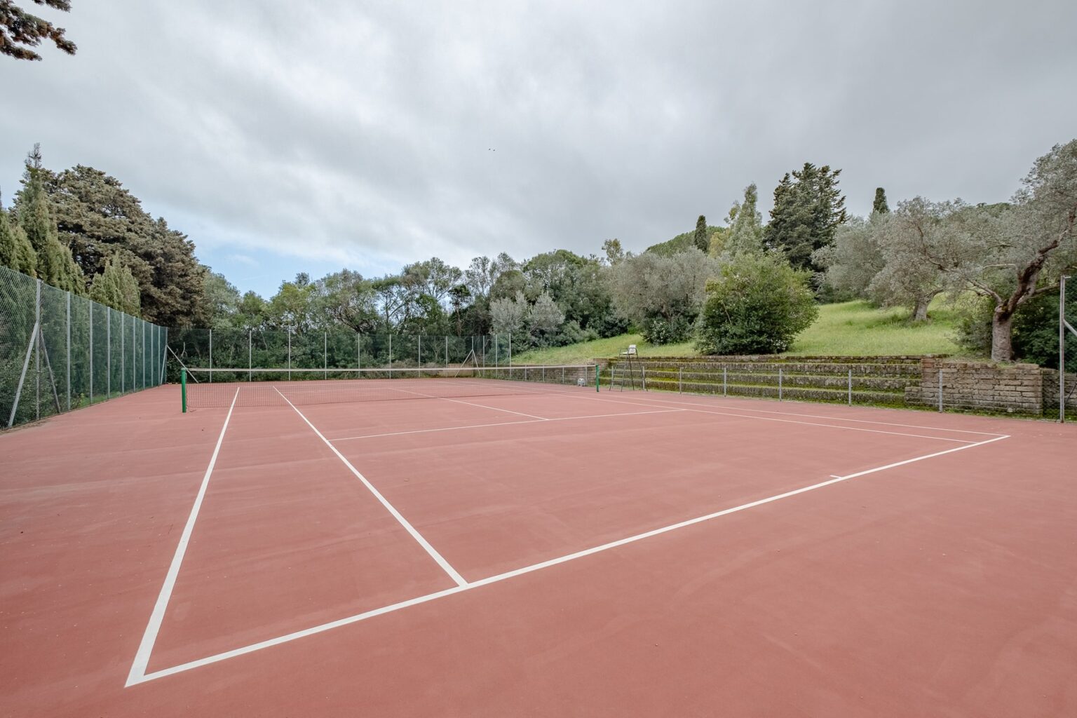 Empty outdoor tennis court with red clay surface surrounded by greenery and trees under a cloudy sky. Ideal for recreational play and training. | Only on SopranoVillas
