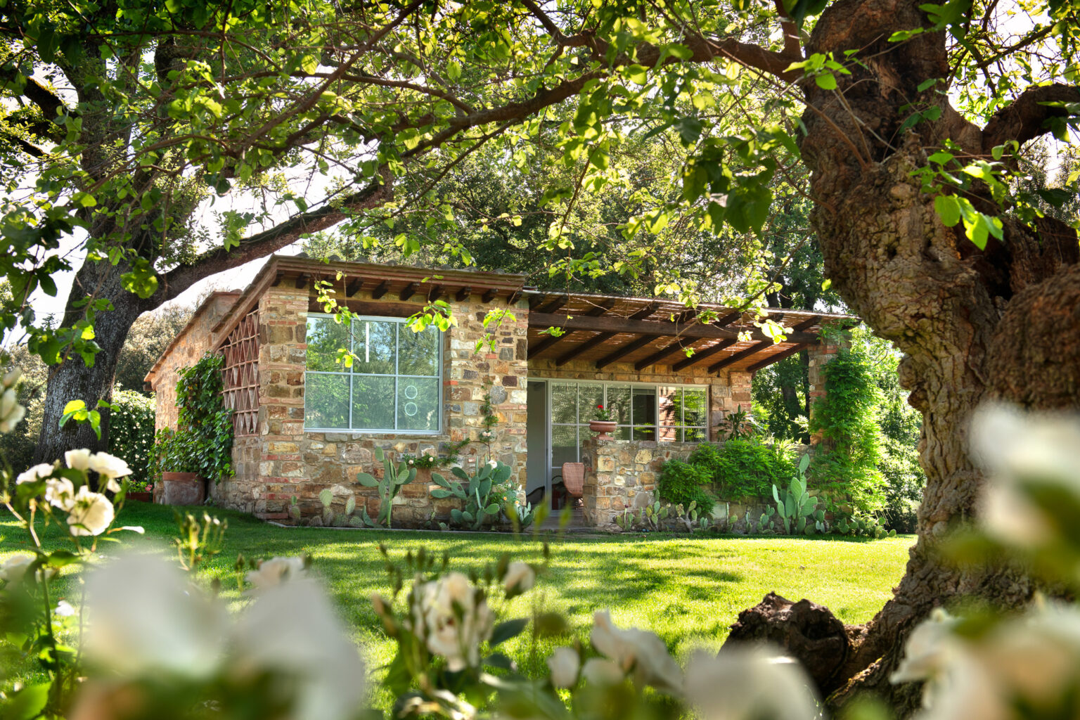Charming stone cottage surrounded by lush greenery and flowers, featuring large windows and a wooden overhang, set in a serene garden environment. | Only on SopranoVillas